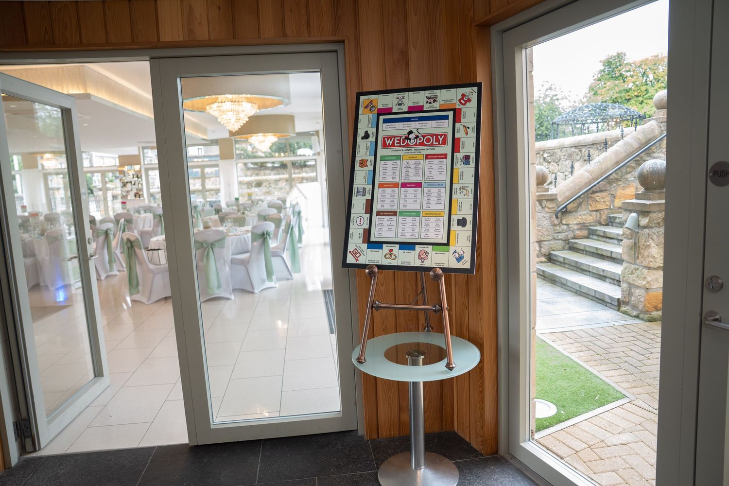 Wedding venue entrance with Monopoly themed seating chart on easel beside glass doors leading to decorated reception hall.