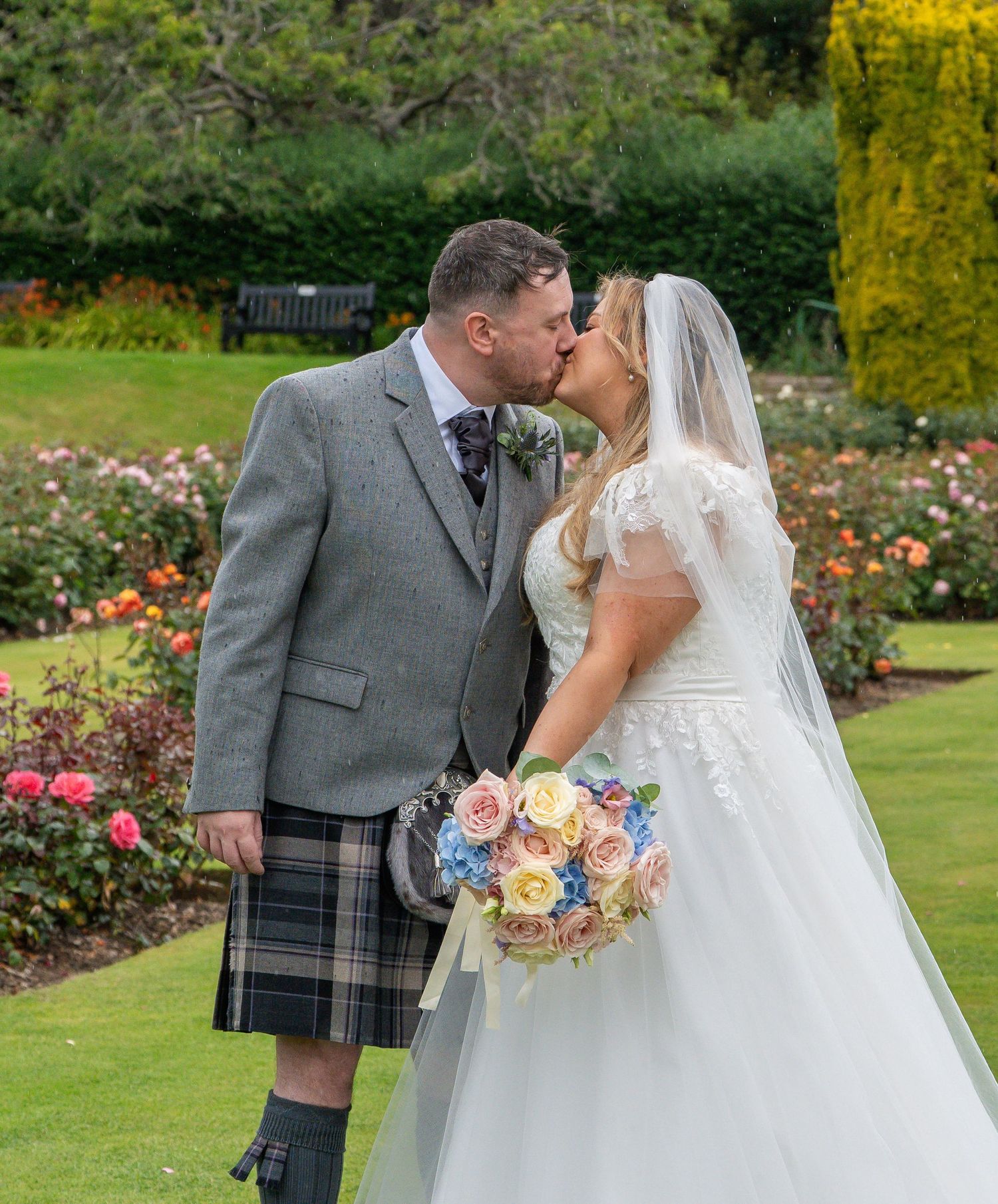 Bride and groom share a romantic kiss in a rose garden, groom wearing traditional Scottish tartan kilt.