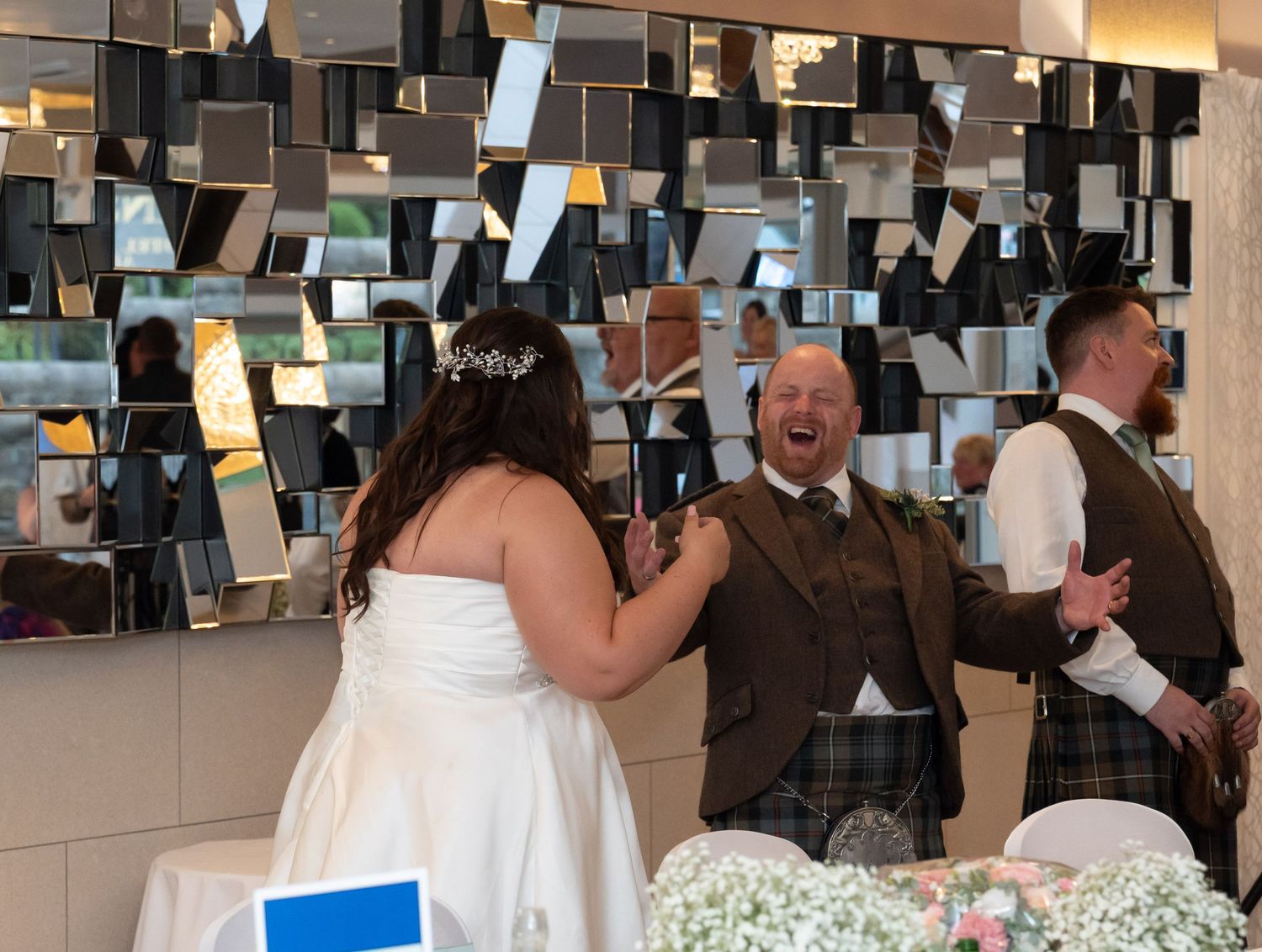 Bride laughing with joyful groom in kilt at wedding reception in front of geometric mirror wall decor.