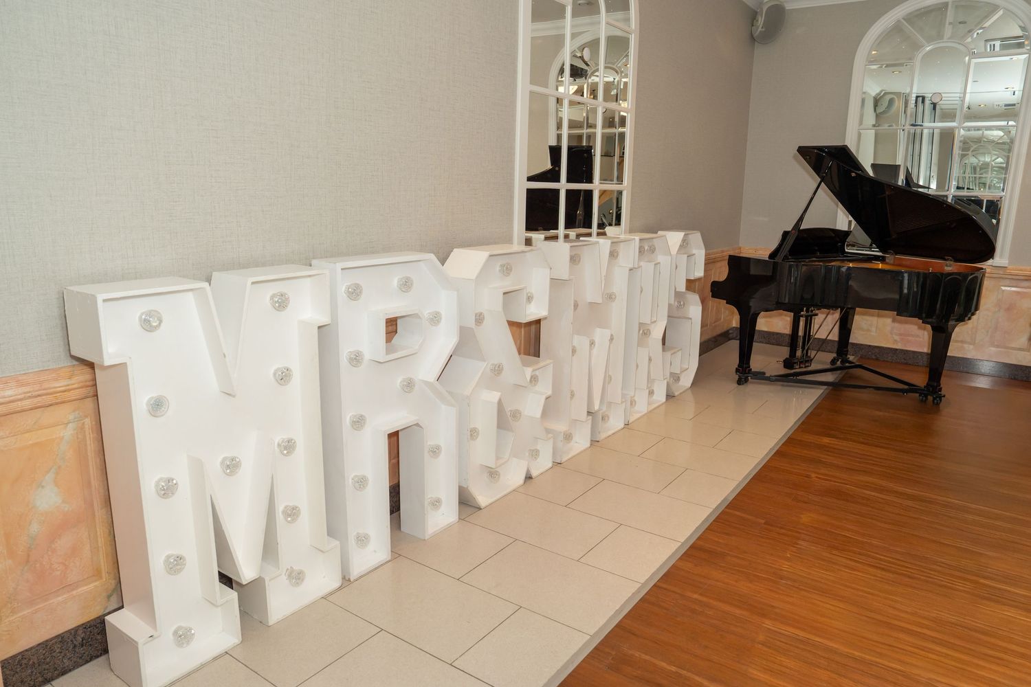 Large white marquee letter lights spelling MRS & MR displayed on a wooden floor beside a grand piano at a wedding venue.