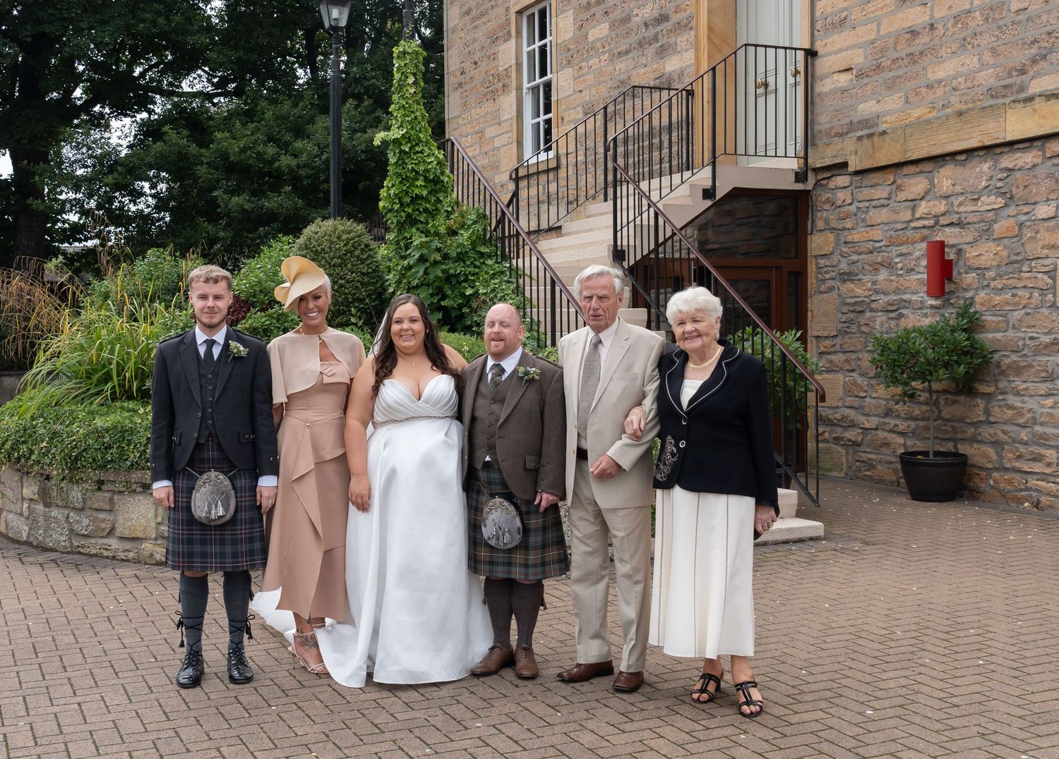 Wedding party of six posing outdoors in Scottish attire, including kilts and formal wear, beside a stone building.