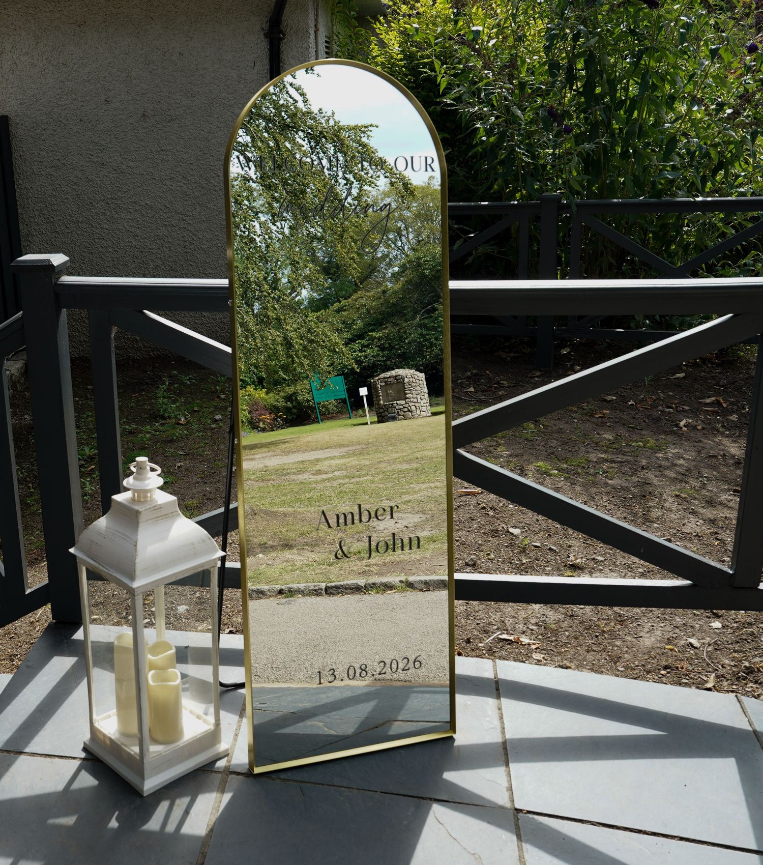 Gold arch wedding welcome mirror sign for Amber and John dated 13.08.2026 displayed outdoors.