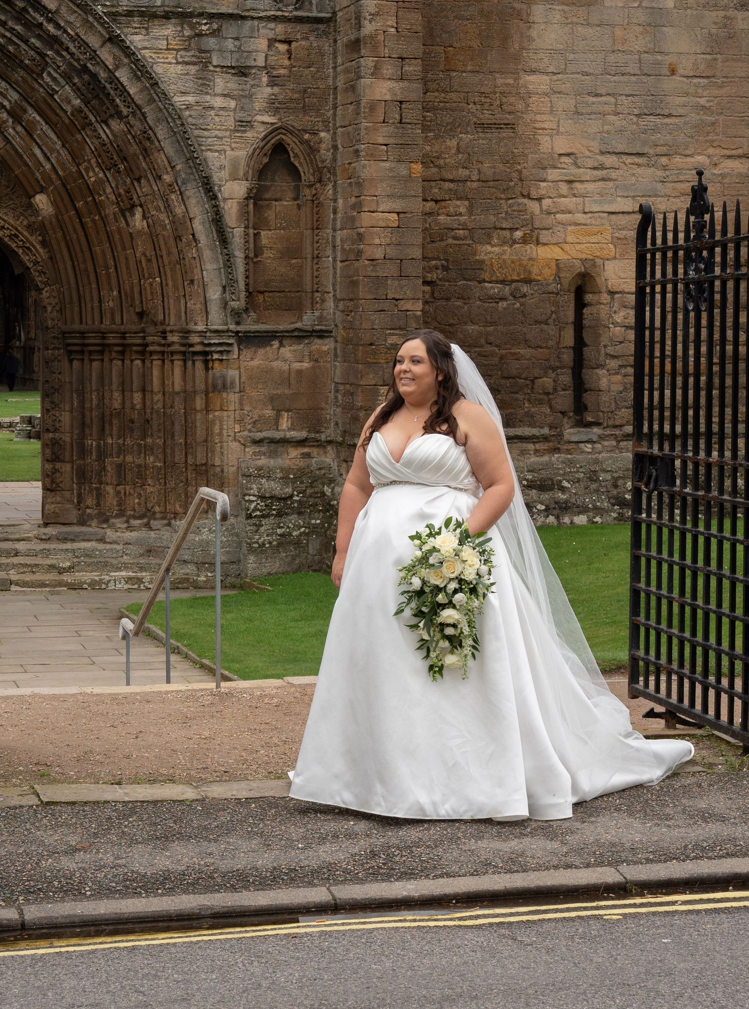 Bride in a white V-neck ball gown with cascading bouquet standing outside historic stone abbey ruins on wedding day.