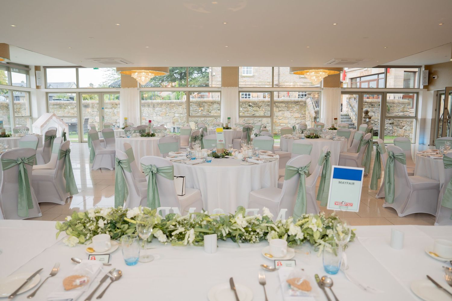 Elegant wedding reception hall with gray chair covers, sage green sashes, round tables, and floral centerpieces in a bright venue.