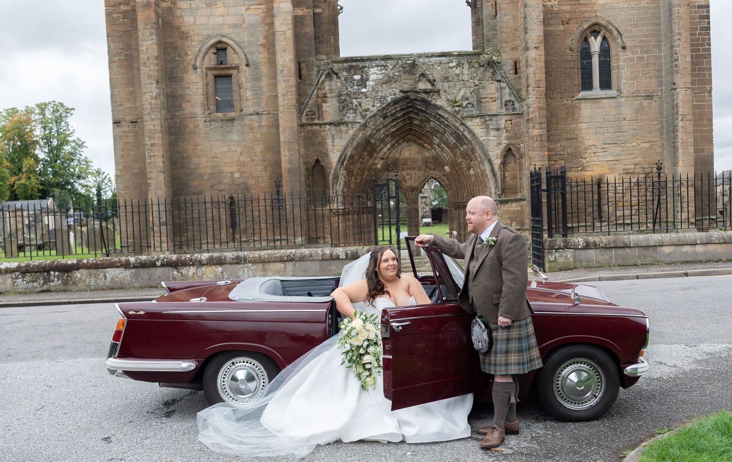 Bride and groom in kilt posing with a vintage burgundy convertible car outside a historic Scottish cathedral.