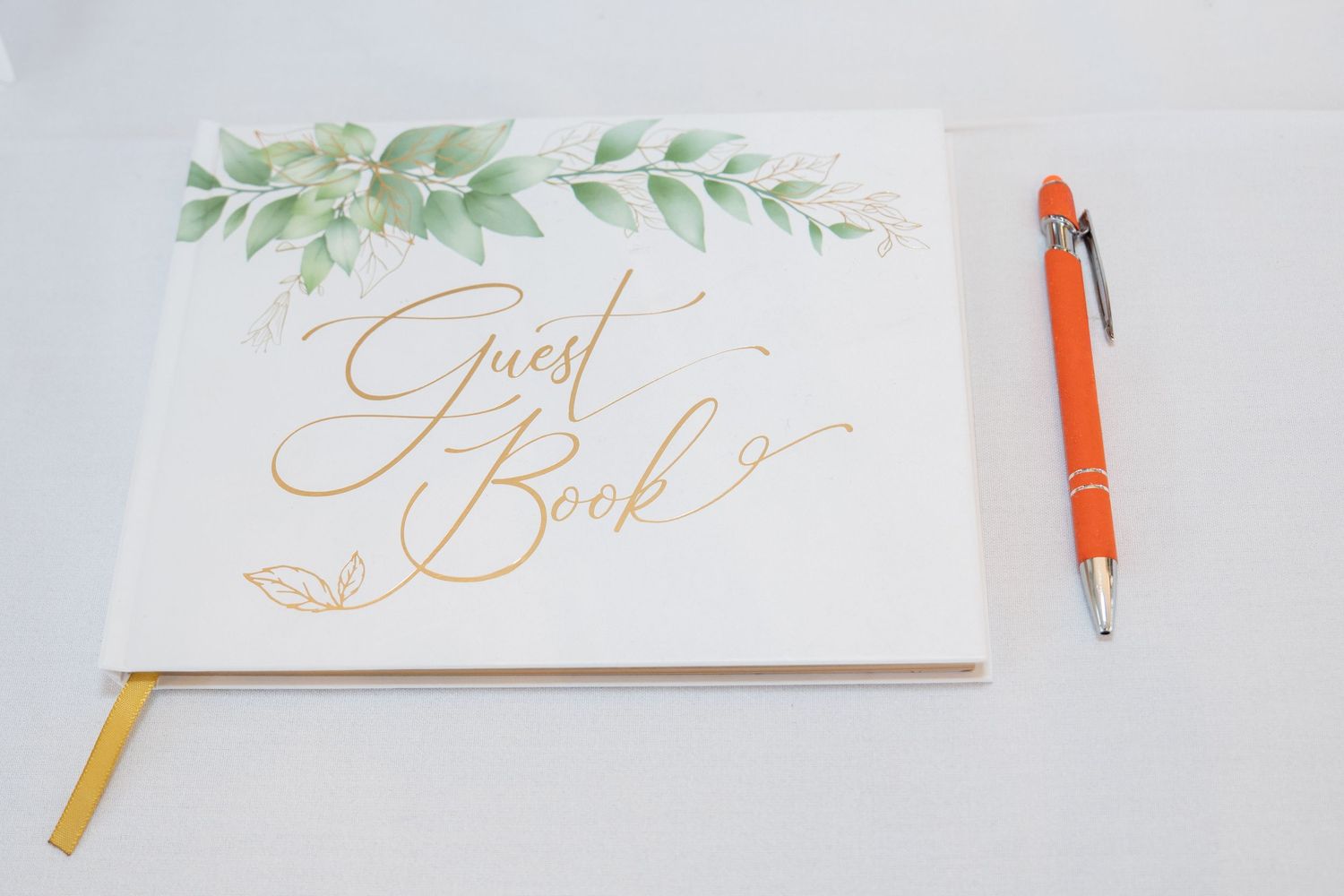 White wedding guest book with gold script lettering and green botanical design, paired with an orange pen.