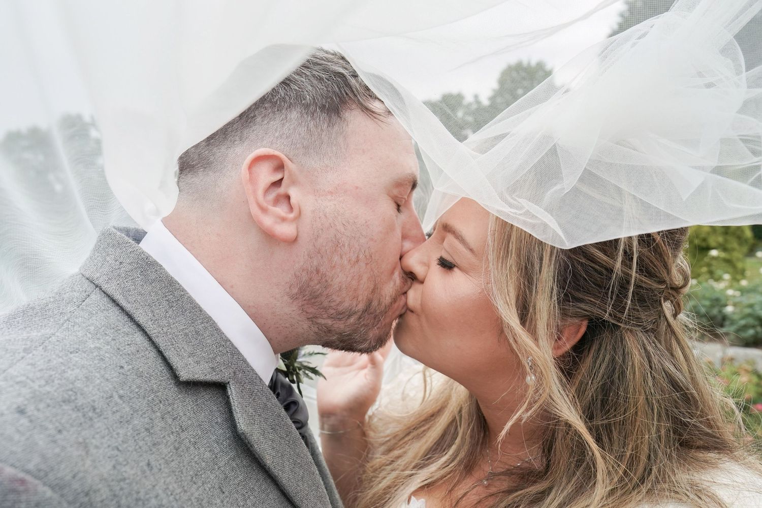Bride and groom share a tender kiss beneath the bride's flowing white veil outdoors.