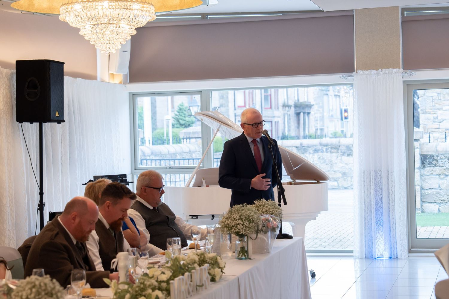 Man delivering a wedding speech at a decorated reception table beneath a crystal chandelier in an elegant venue.