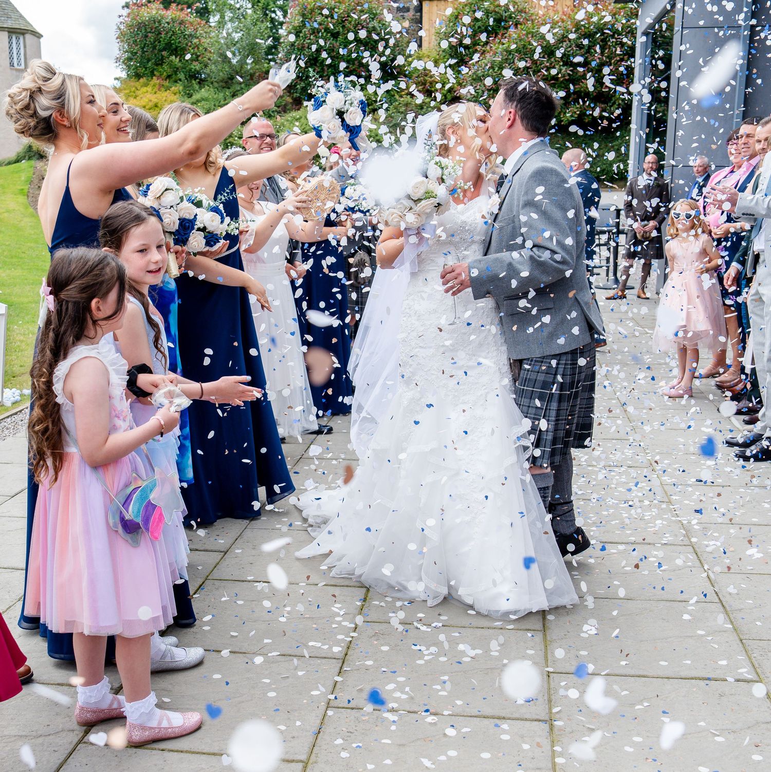 Bride and groom walk through confetti shower surrounded by guests at outdoor wedding celebration.