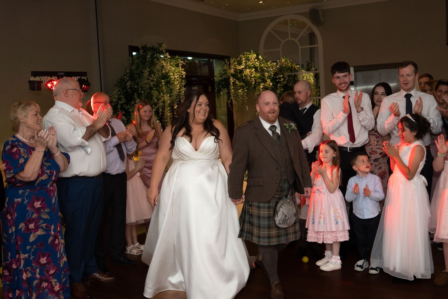 Bride and groom in kilt making grand entrance to wedding reception with cheering guests lining the aisle.