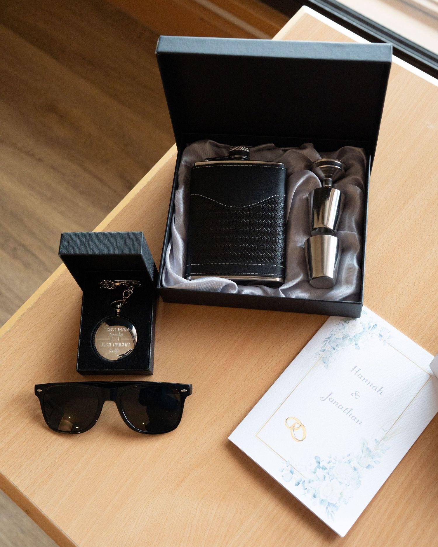 Groomsmen gift set featuring a black leather hip flask, shot glasses, sunglasses, pocket watch, and wedding card.