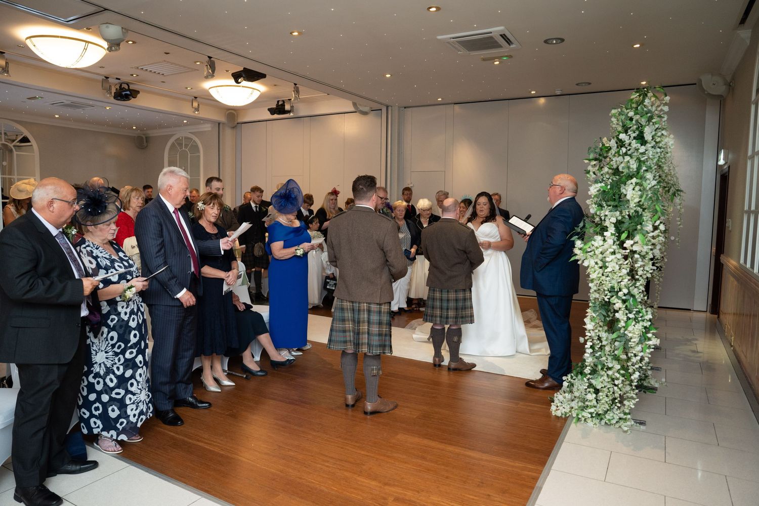Wedding ceremony guests gathered around couple at floral arch in elegant modern wedding venue.