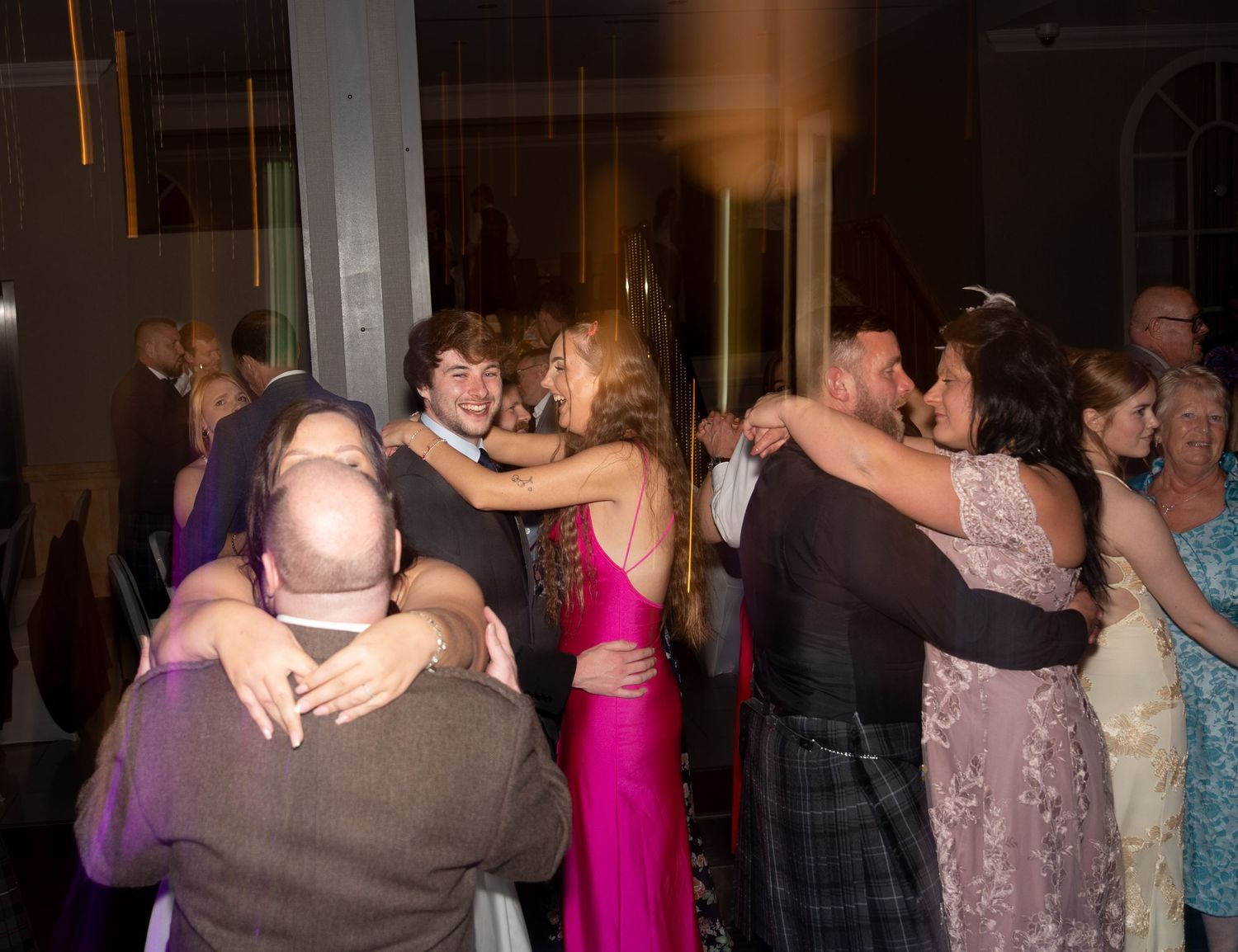 Couples dancing and embracing at a formal event, with guests in elegant attire including kilts and gowns.