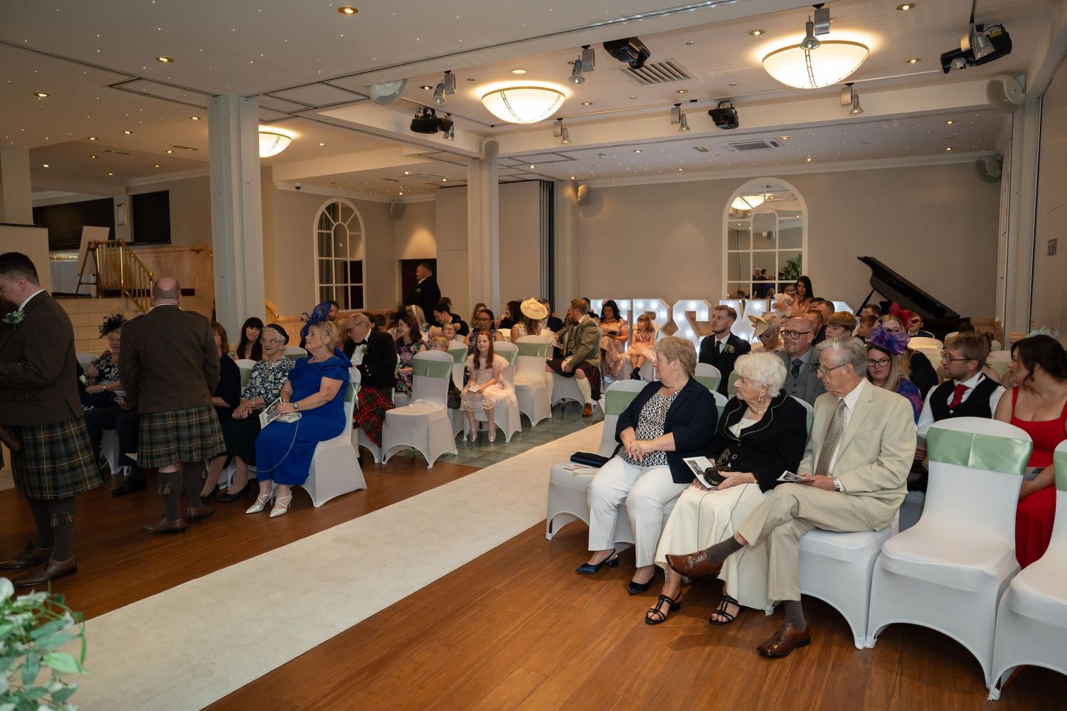 Guests seated in elegant white-decorated wedding venue hall during Scottish wedding ceremony.