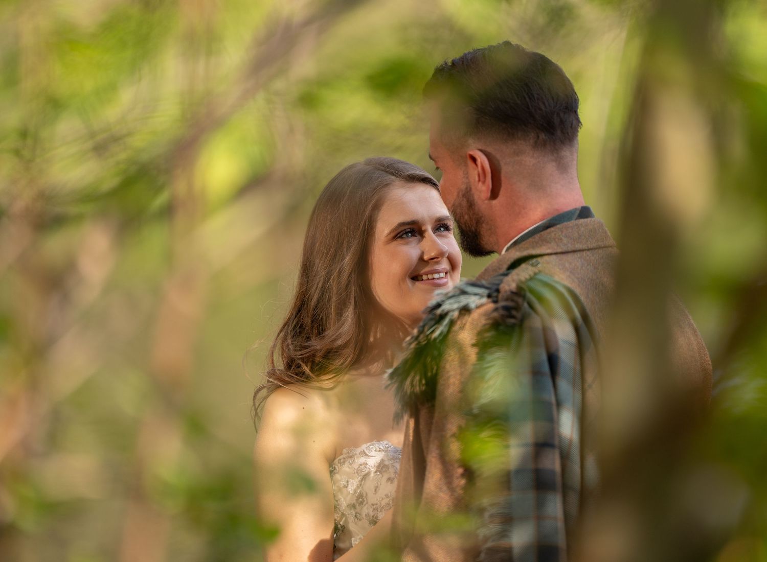 A smiling bride gazes at her groom surrounded by lush green trees in a romantic outdoor wedding portrait.