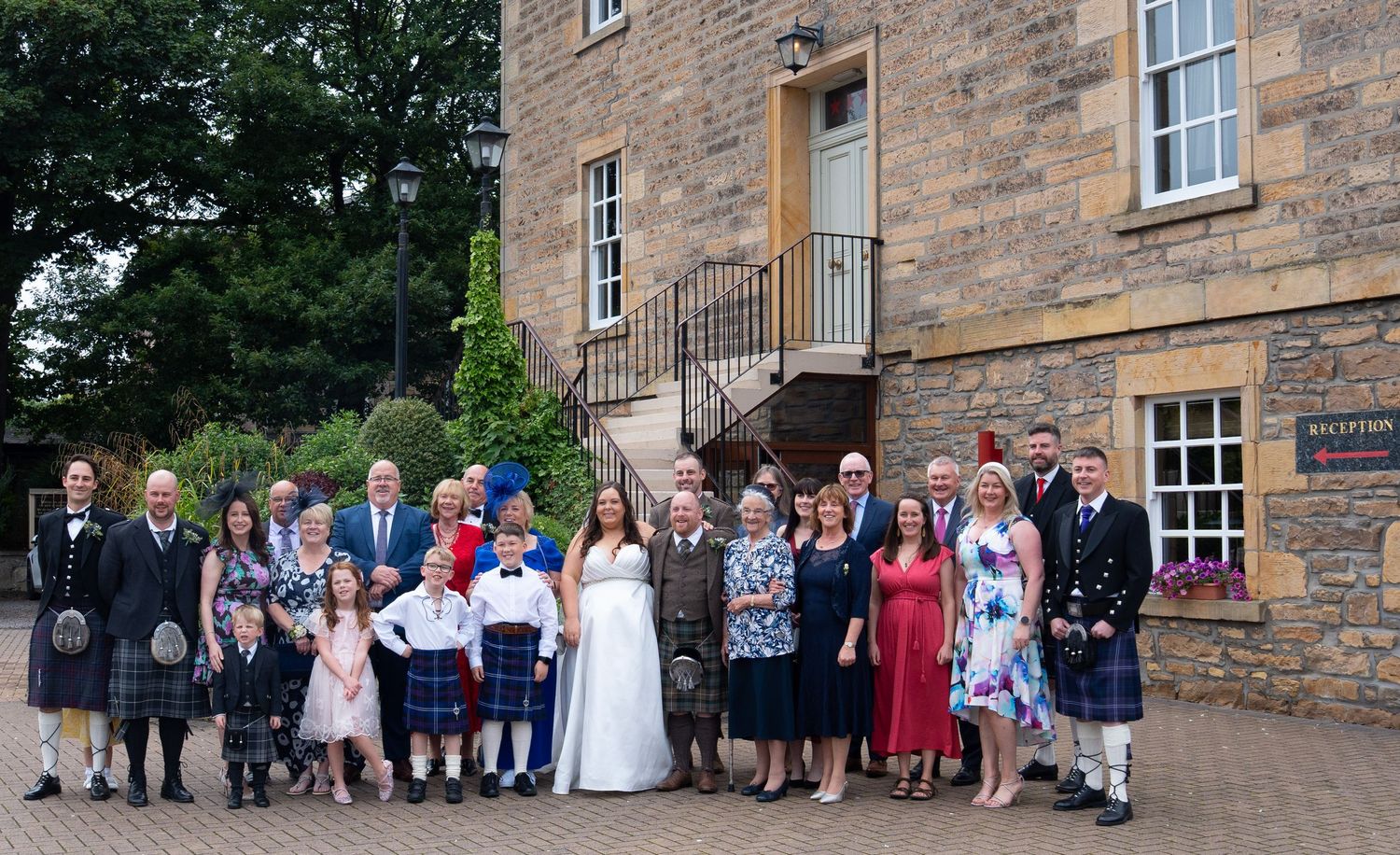Wedding party group photo outside a Scottish stone building with guests in kilts and formal attire.