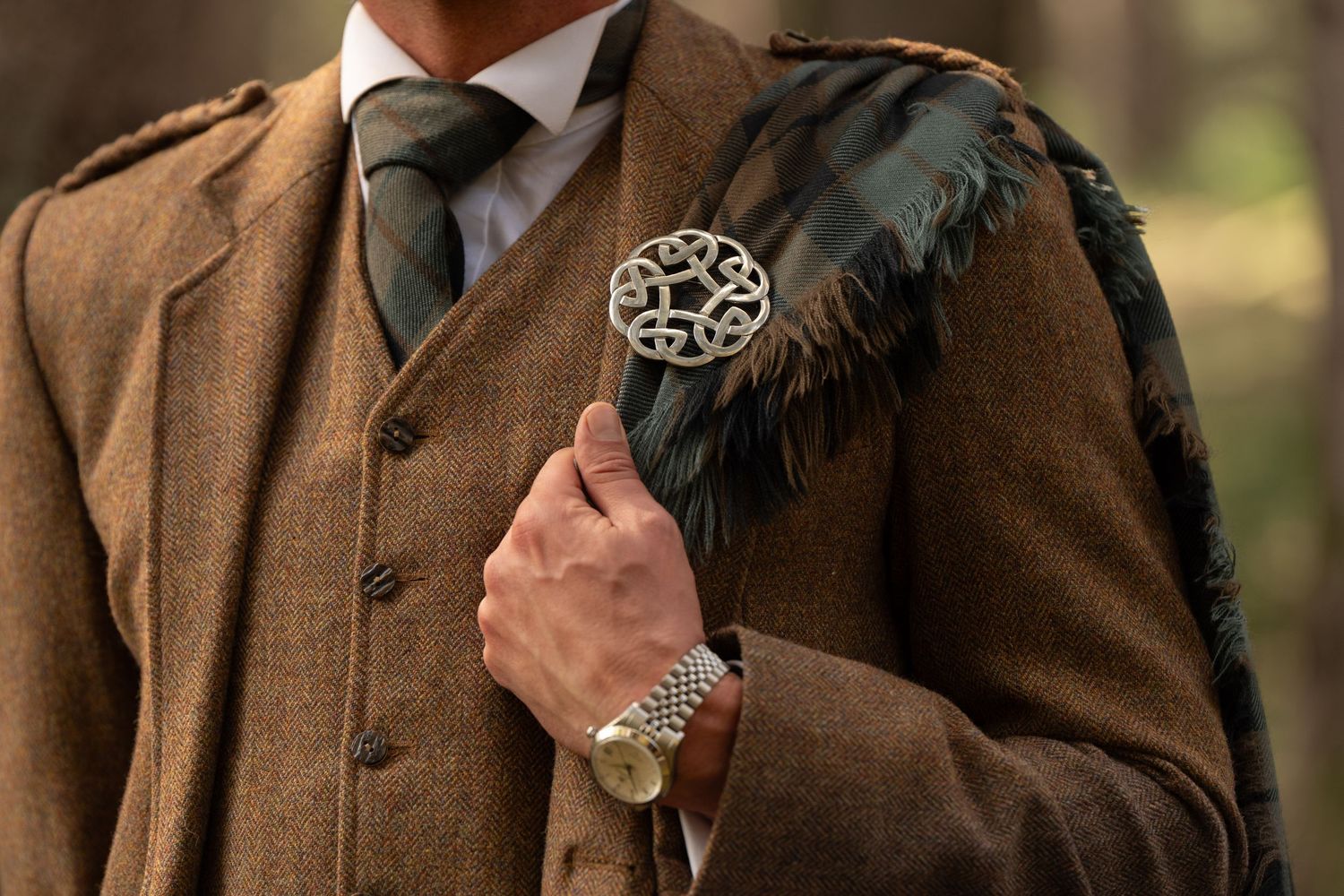 Man in brown tweed jacket with Celtic brooch pin and green tartan pocket square, wearing a silver watch.