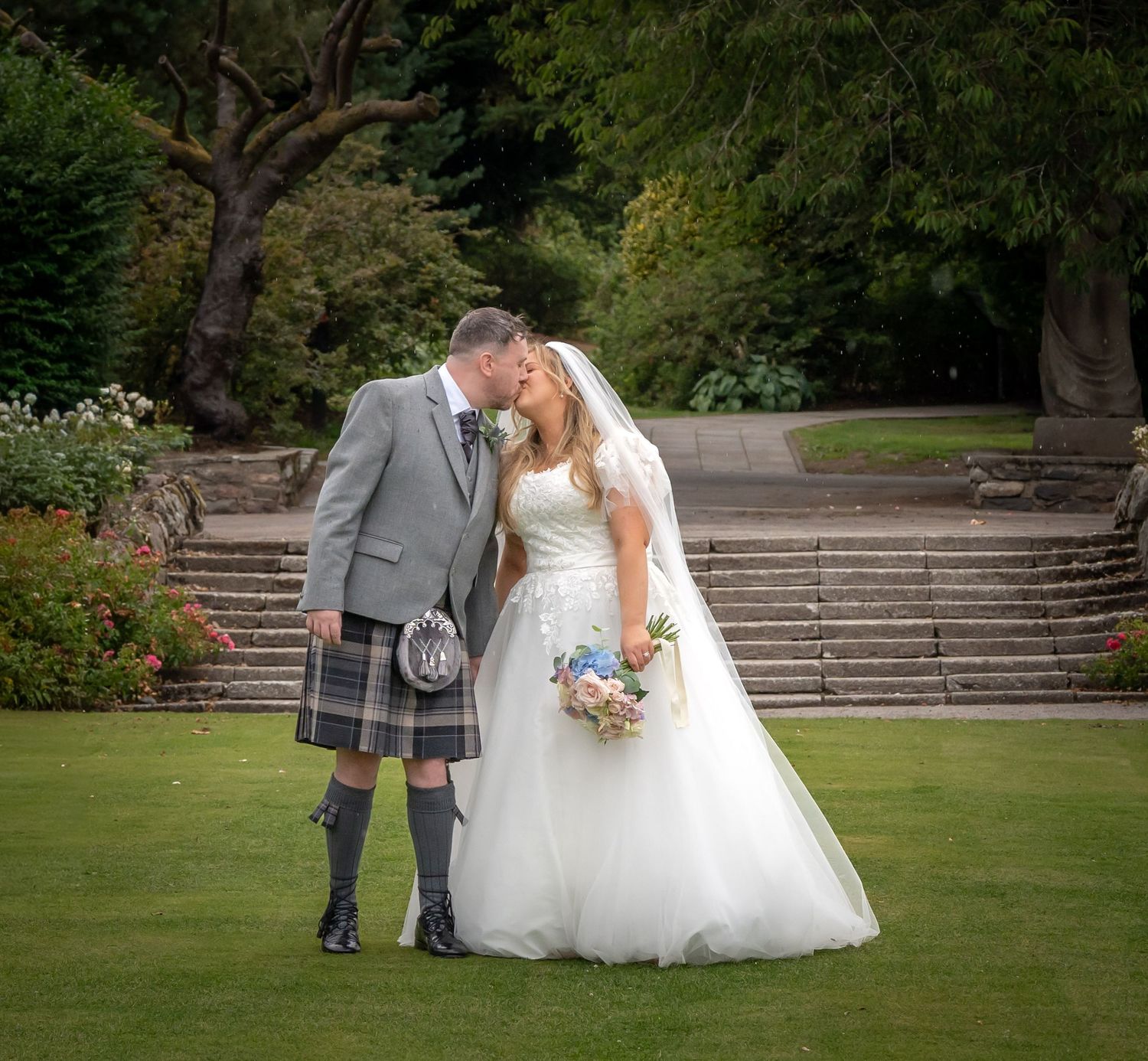 Bride and groom share romantic kiss in garden, groom wearing traditional Scottish kilt.