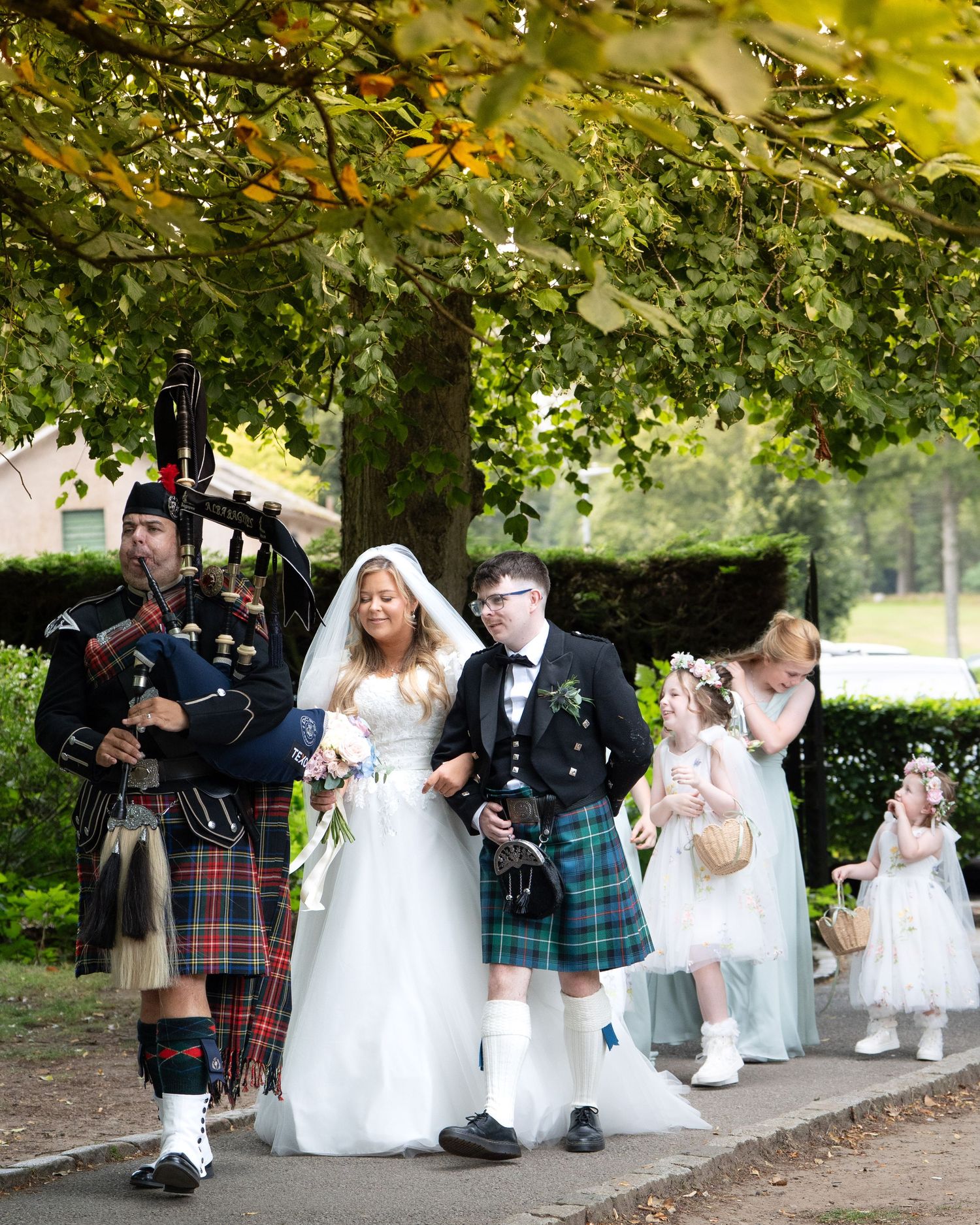 Bagpiper leads bride, groom in tartan kilt, and flower girls in outdoor wedding procession.