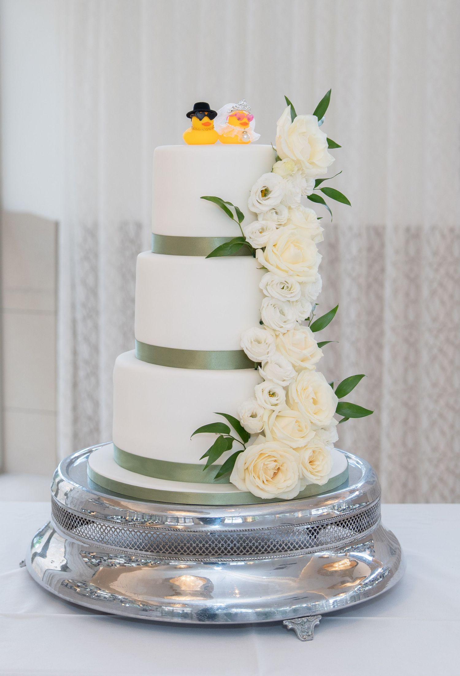 Four-tier white wedding cake with sage green ribbon, cascading white roses, and rubber duck bride and groom cake toppers.