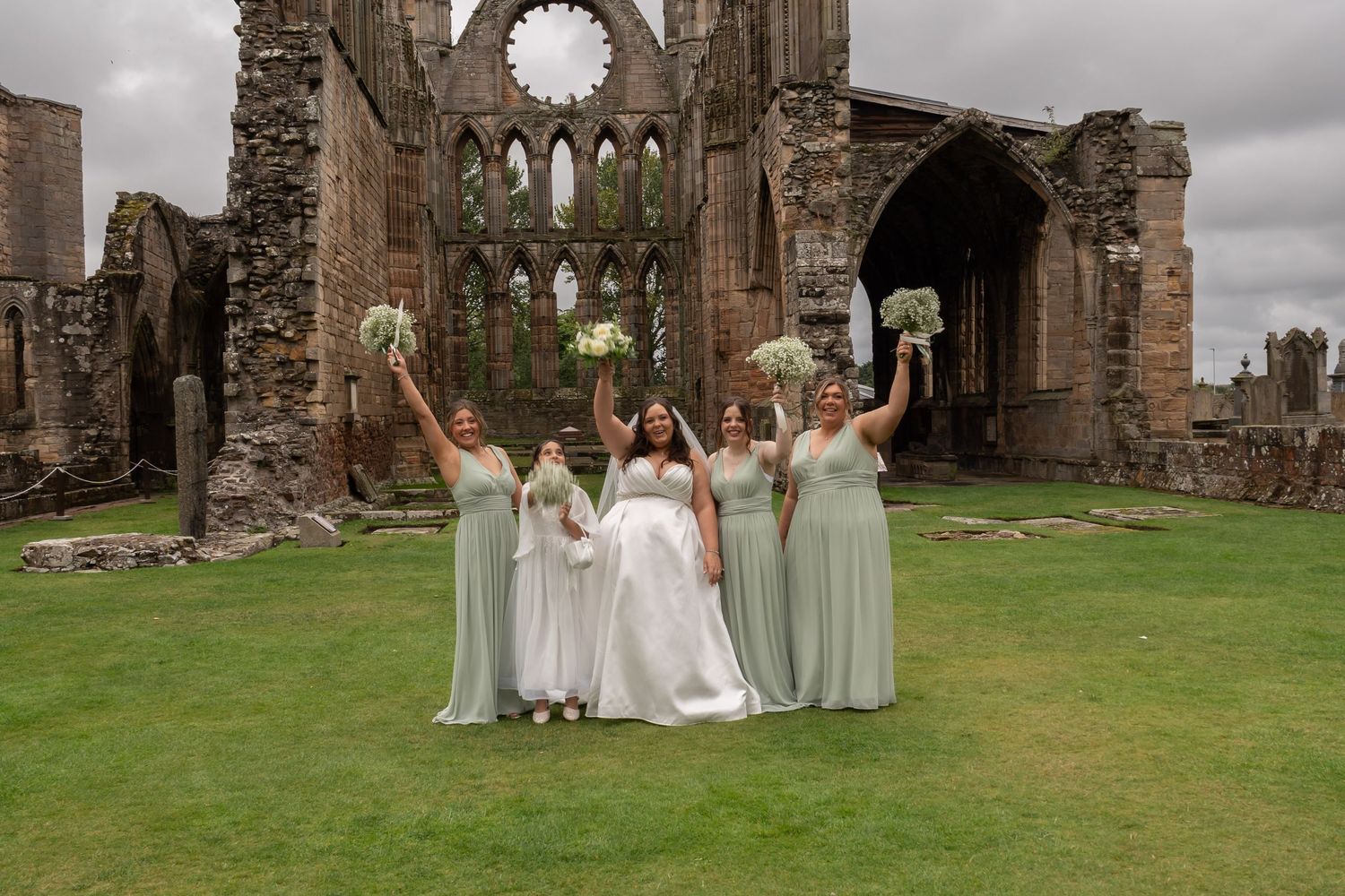Bride and four bridesmaids in sage green dresses celebrate with bouquets raised at historic stone abbey ruins.