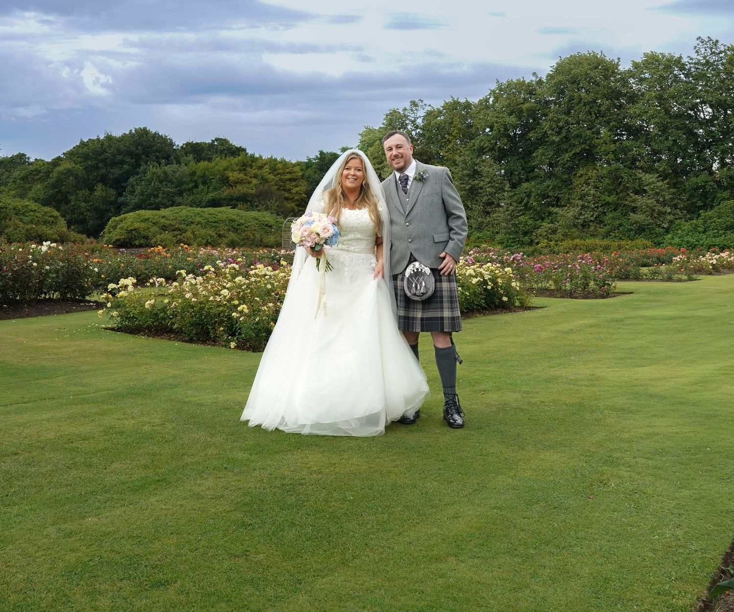 Bride in white gown and groom in traditional Scottish kilt pose together on lush green lawn surrounded by rose gardens.