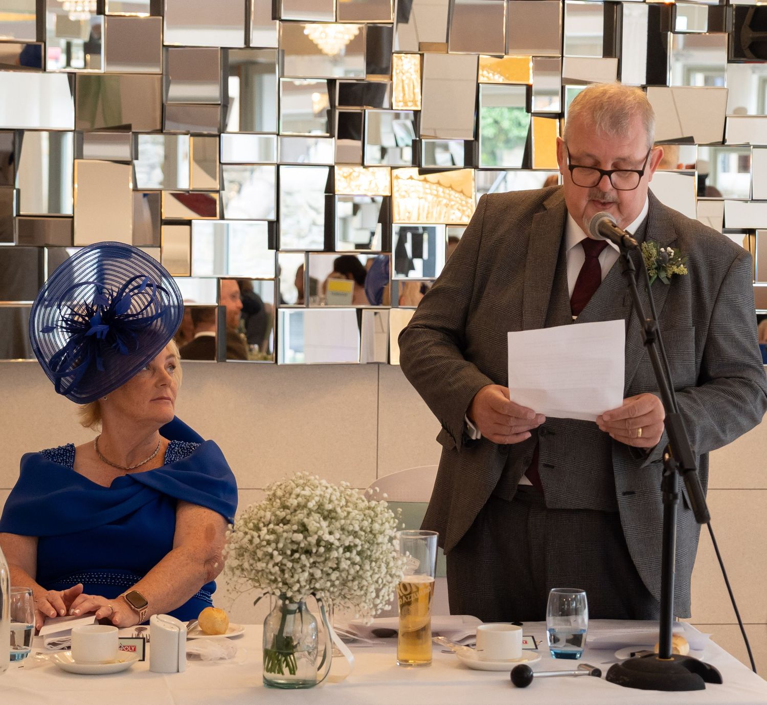 Father of bride giving wedding speech at microphone beside woman in blue dress and fascinator hat.