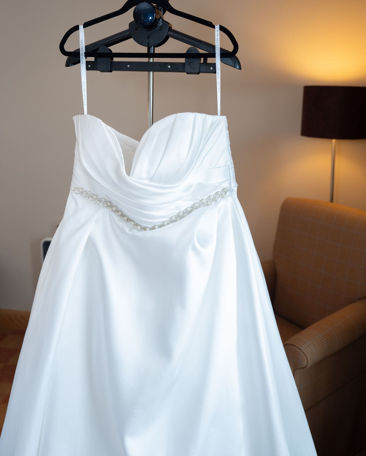 Strapless white satin wedding dress with beaded belt hanging on a hanger.