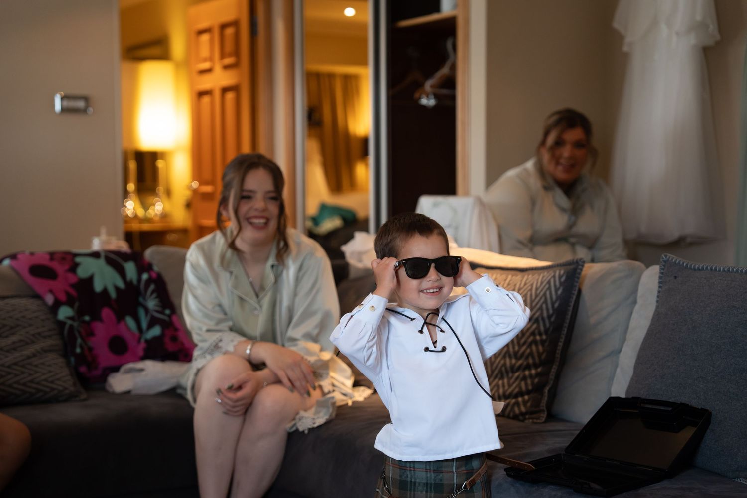 A young boy wearing sunglasses sits on a couch smiling with two women in a warmly lit room.
