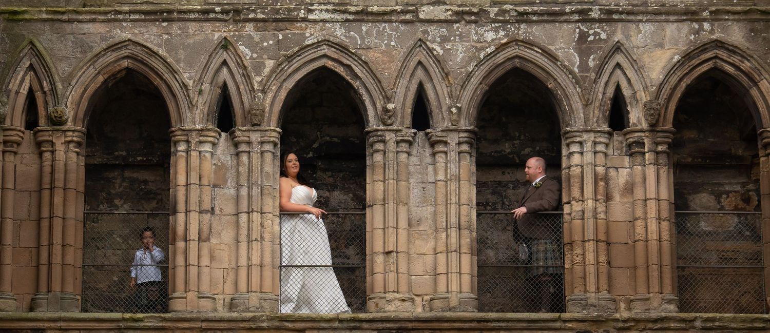 Bride and groom posing in Gothic stone arches of a historic abbey, with a photographer visible in the background.