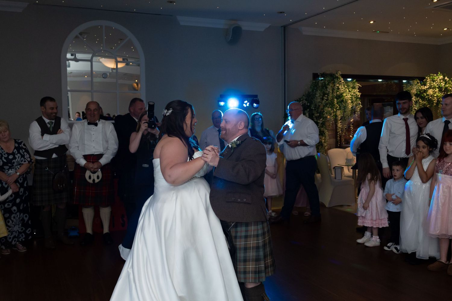 Bride and groom sharing romantic first dance under blue lighting with wedding guests watching in the background.
