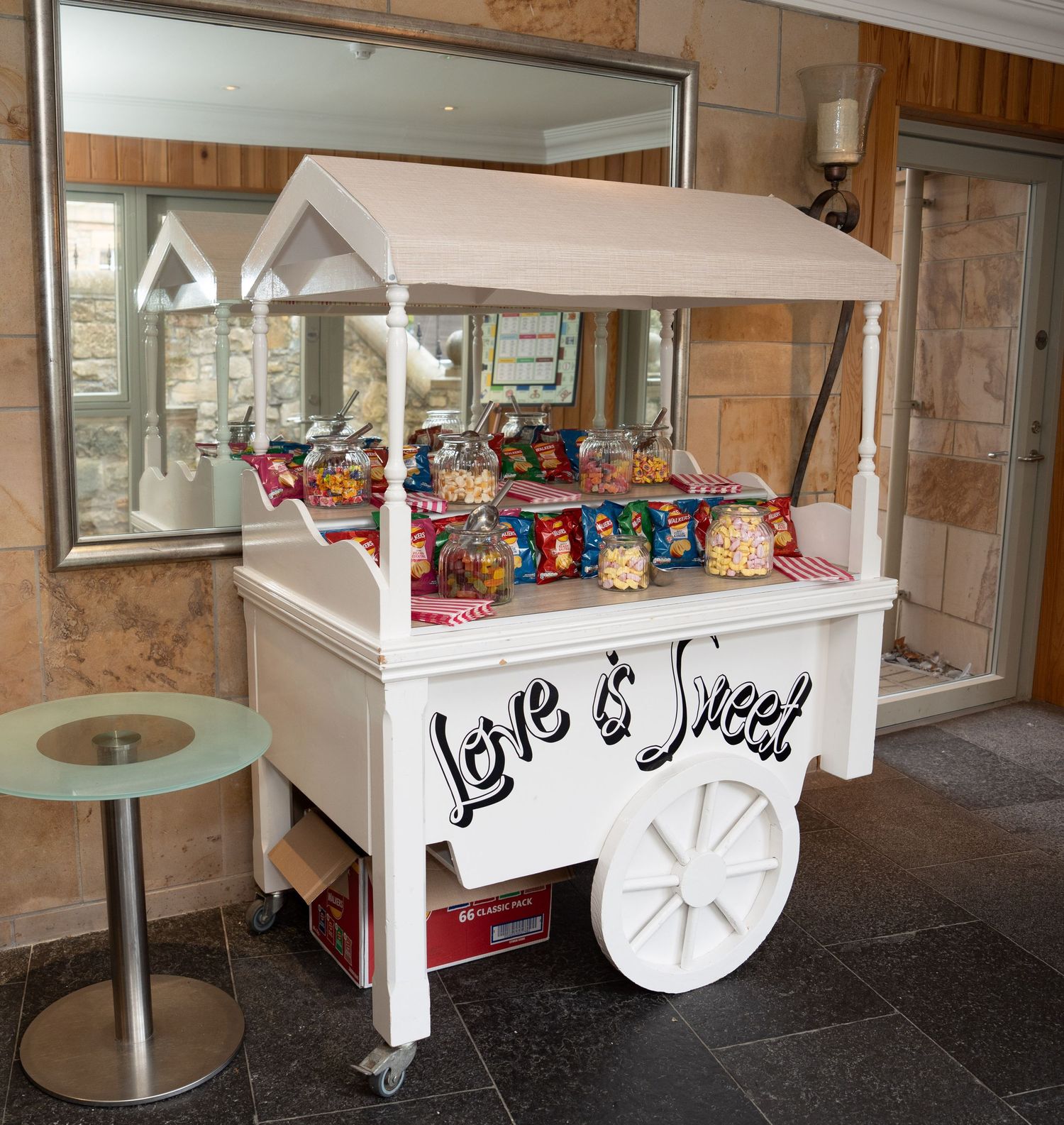 White wedding candy cart with Love is Sweet sign displaying colorful sweets and snacks at a wedding venue.