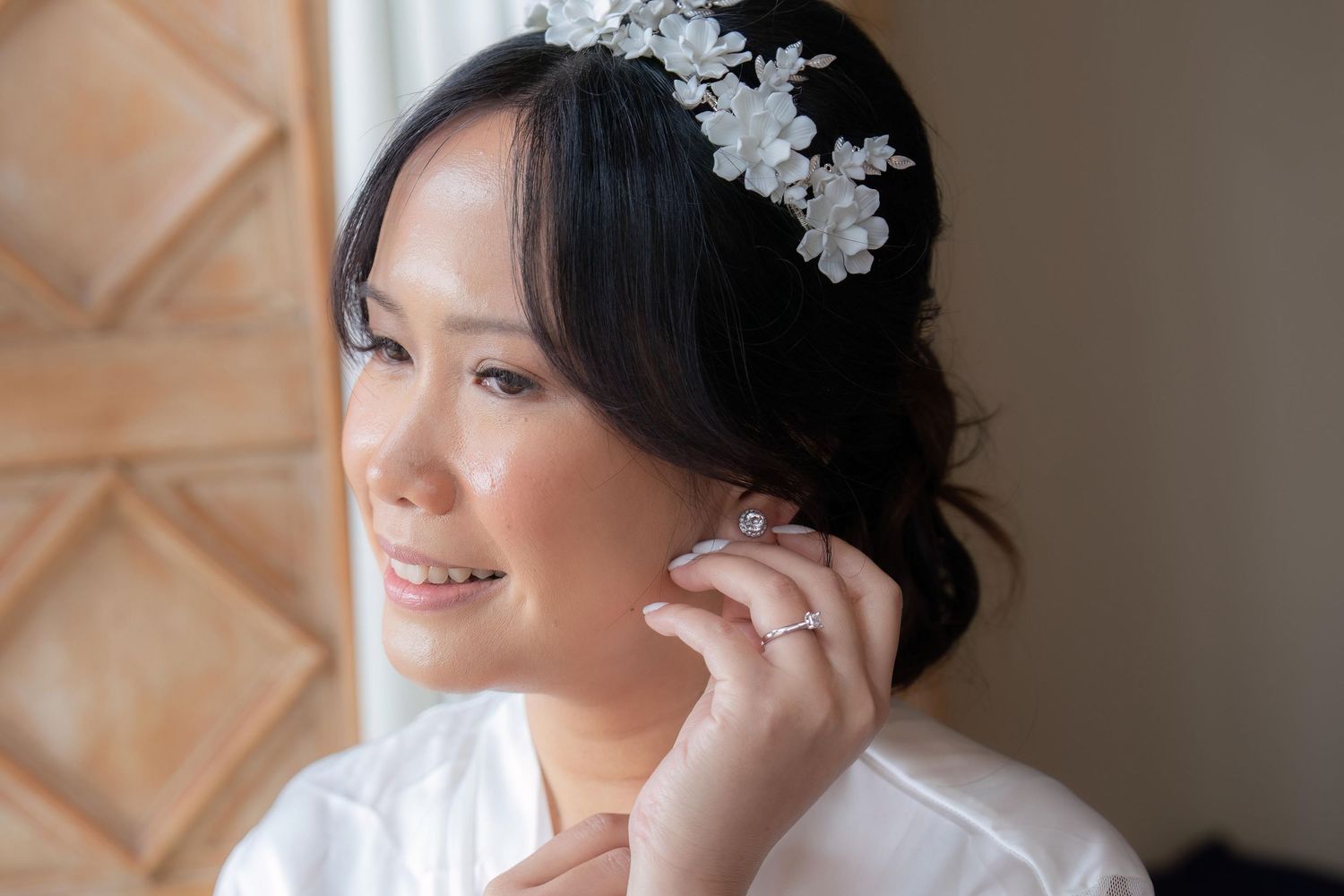 A bride wearing a floral headpiece adjusts her earring while smiling softly.
