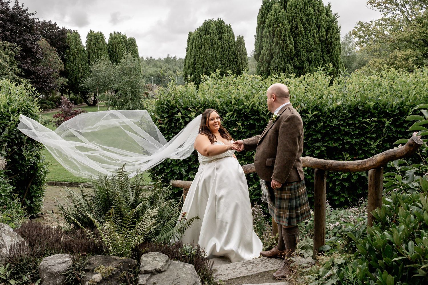 Bride and groom share a romantic moment in lush garden as her veil billows in the wind on their wedding day.