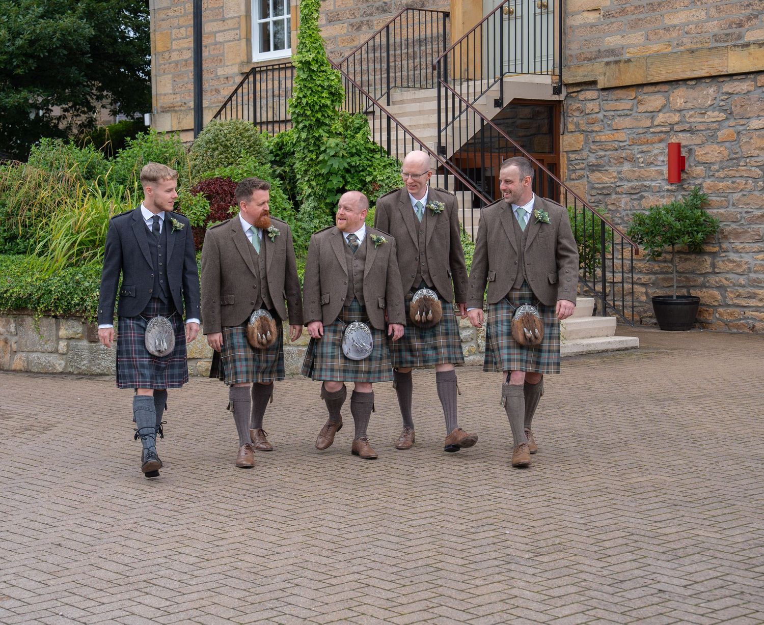 Five groomsmen in traditional Scottish kilts and tweed jackets walking outside a stone wedding venue.