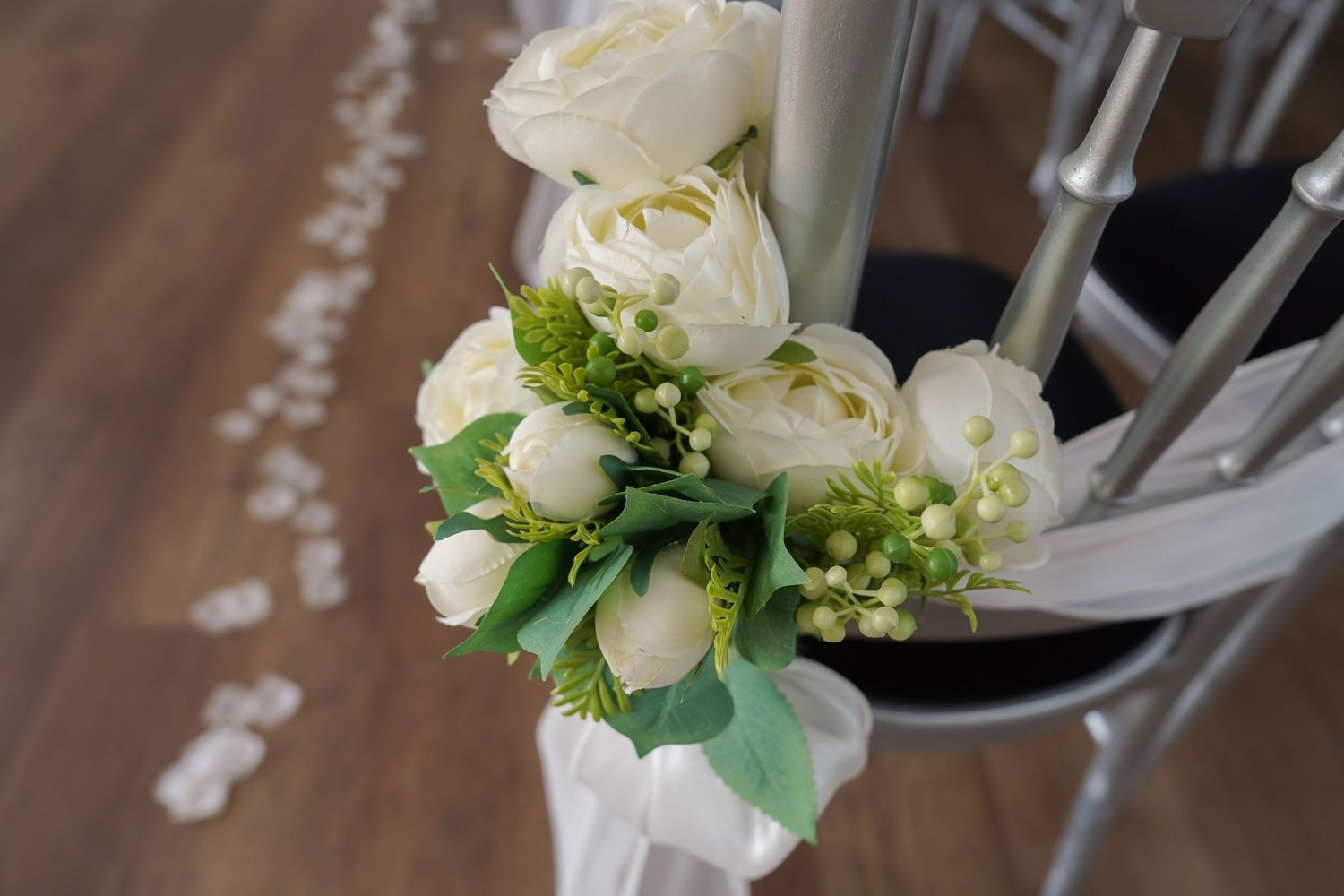 Elegant white ranunculus bouquet tied with ribbon decorates wedding ceremony chair aisle.
