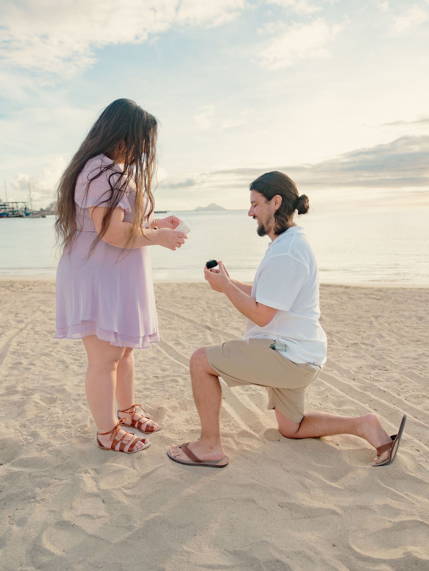 Man proposing on one knee to woman in lavender dress on sandy beach at sunset.