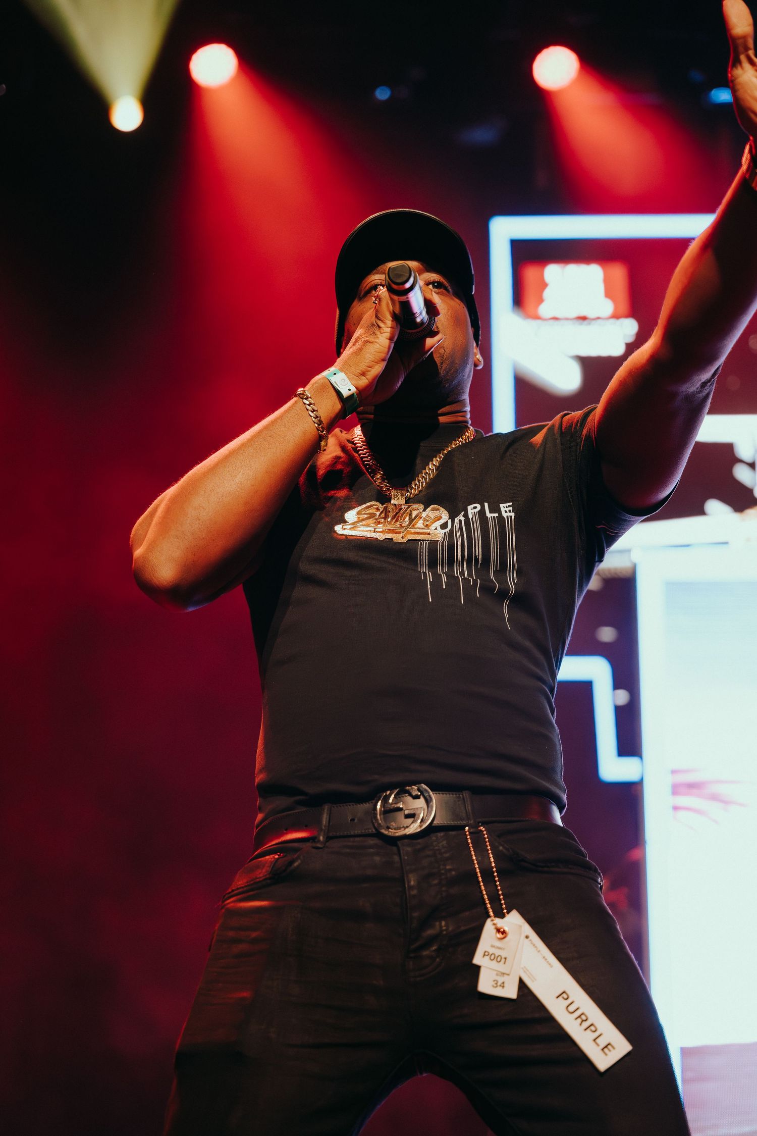 Male artiste performs on stage wearing black cap and gold chain under red stage lights.