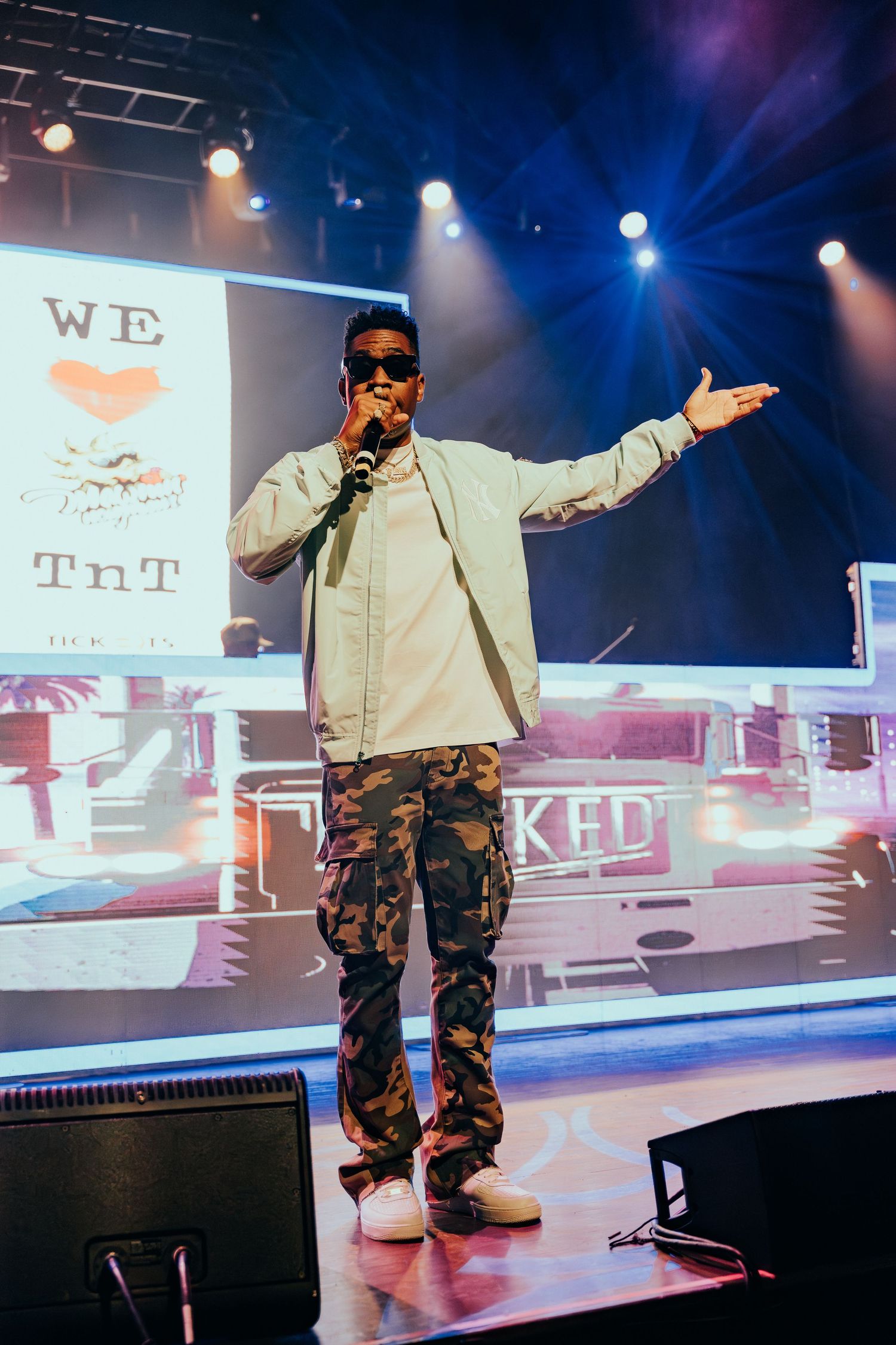 Male artiste in camo pants and light jacket performing on stage at a live concert.