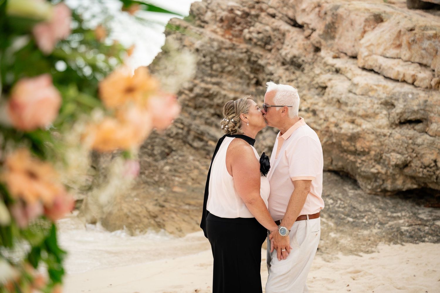 Older couple sharing a kiss on sandy beach framed by peach floral arrangement during vow renewal.