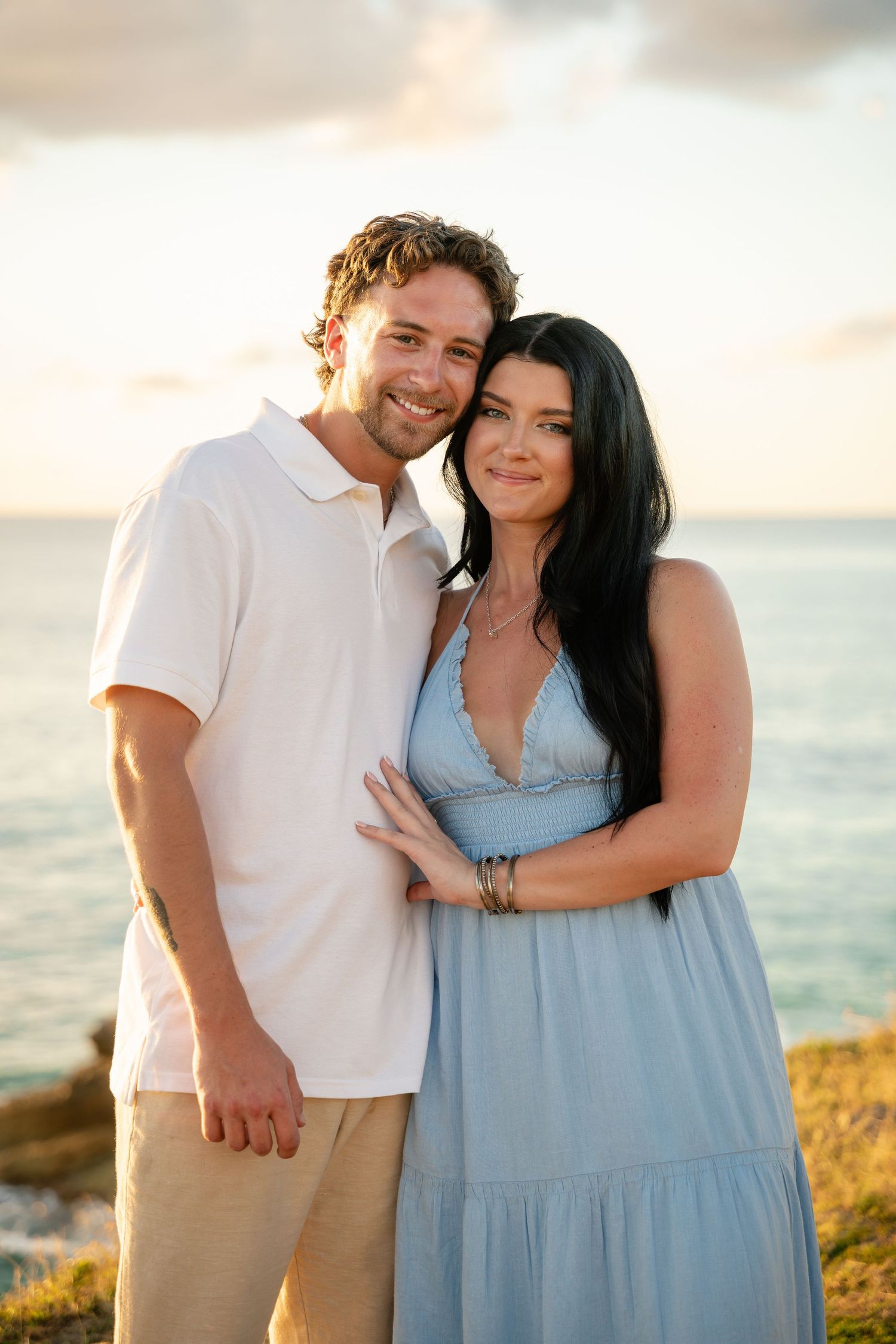 Smiling couple posing at golden hour by the ocean, man in white polo and woman in blue dress with dark hair.