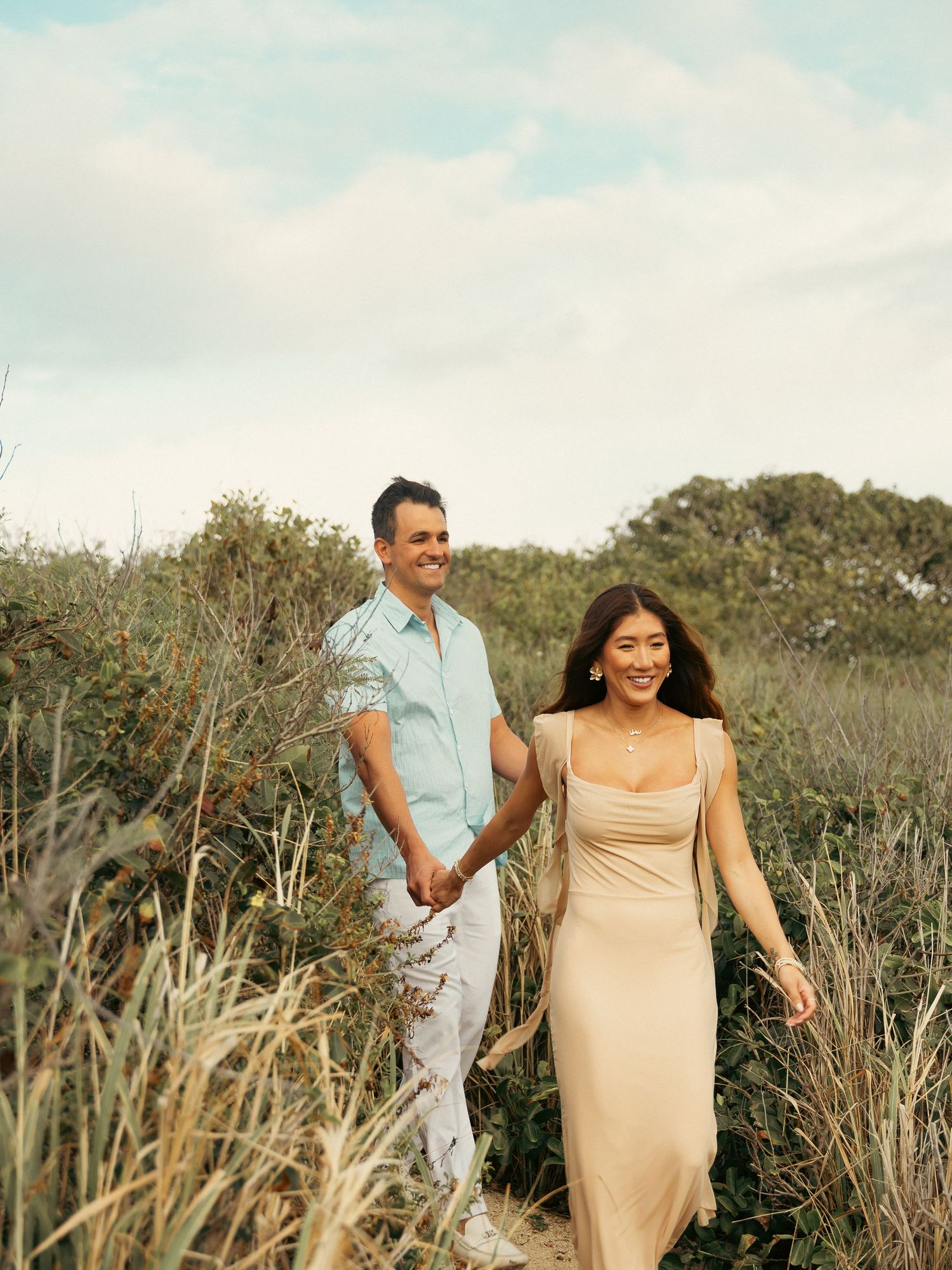 A smiling couple holds hands while walking through tall coastal grass and dunes on a cloudy day.