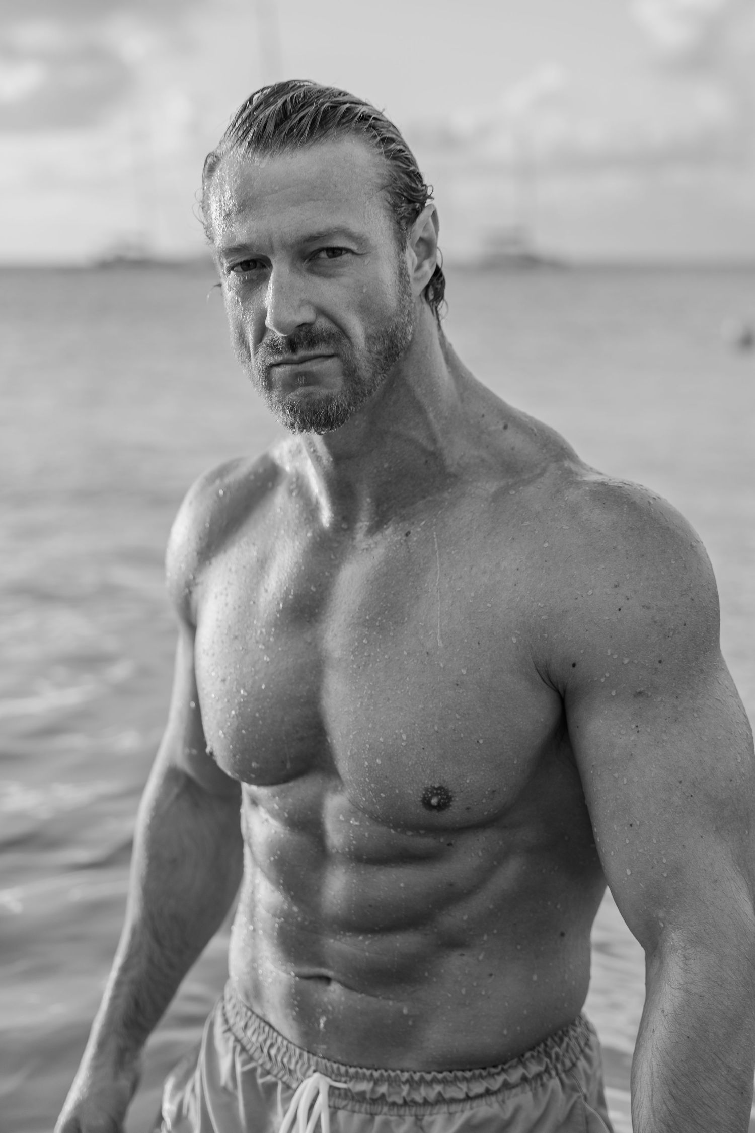 Black and white portrait of a muscular shirtless man standing at the beach.