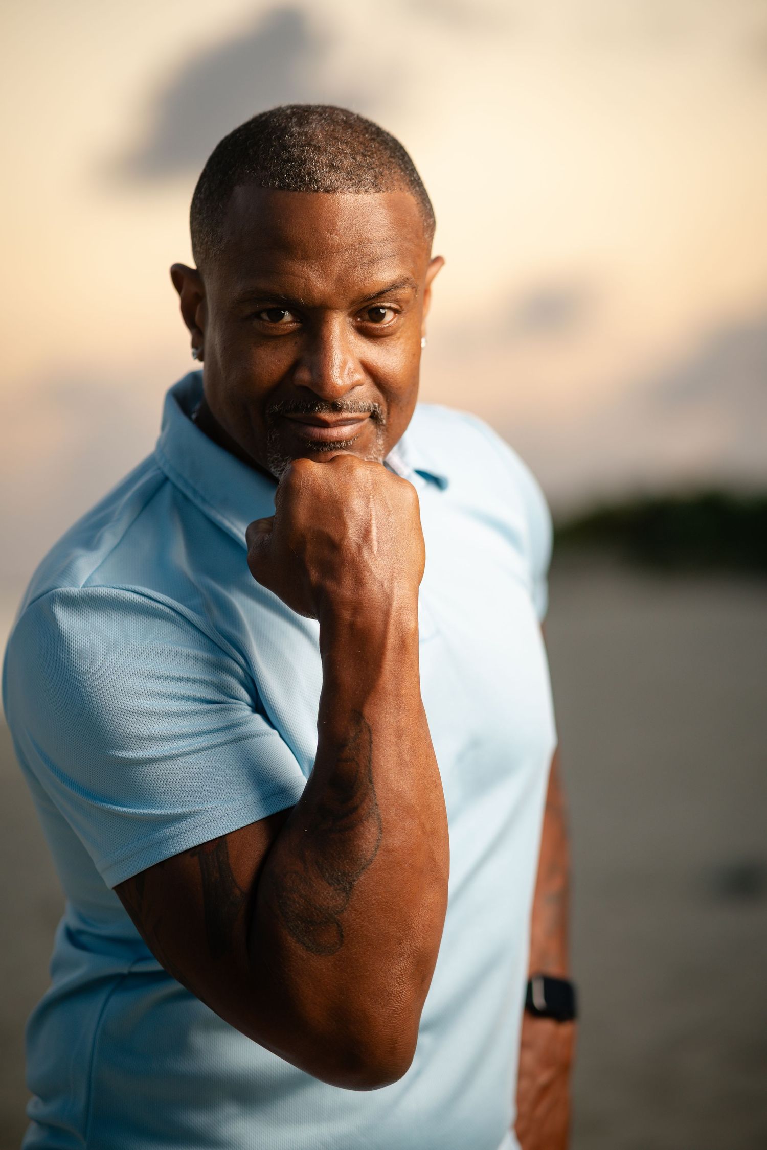 Confident Black man in a light blue polo shirt posing with a raised fist at sunset on a beach.