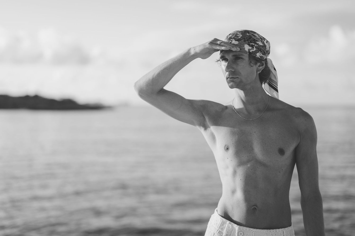 Black and white photo of shirtless man shielding eyes gazing at ocean horizon at sunset.