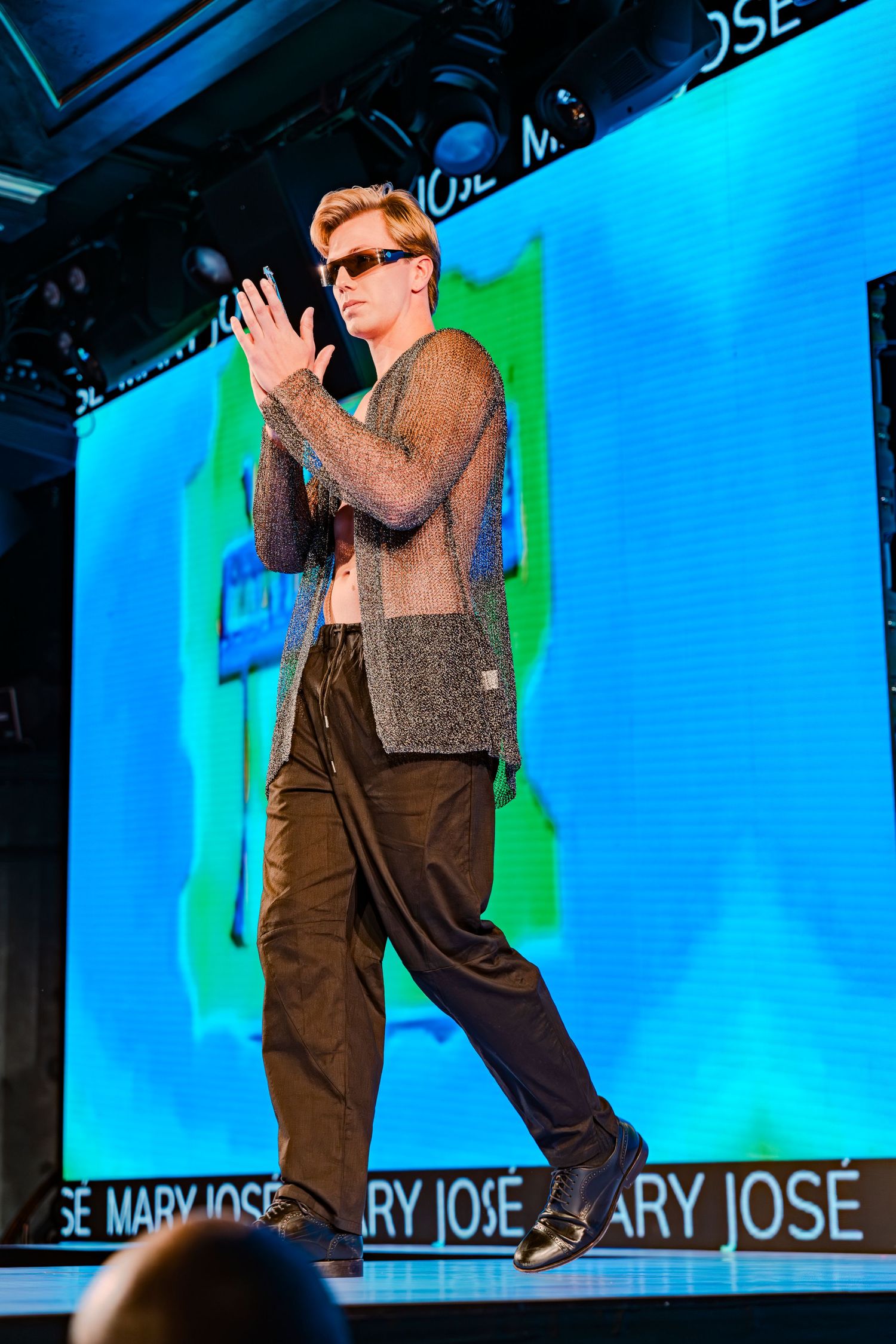 Male model walks runway in sheer mesh top, dark trousers, and sunglasses at Mary José fashion show.