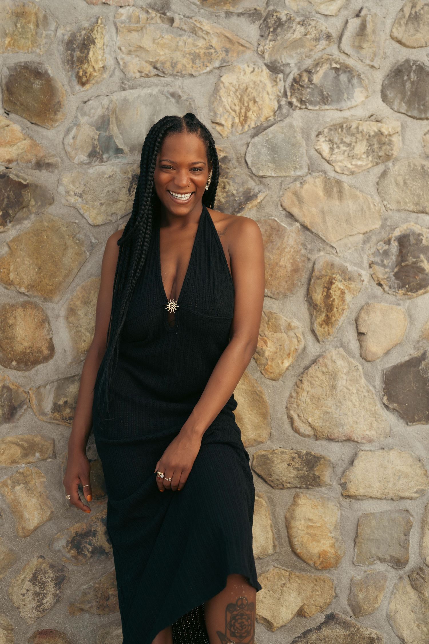 Smiling woman in black halter dress with braids leaning against stone wall outdoors.