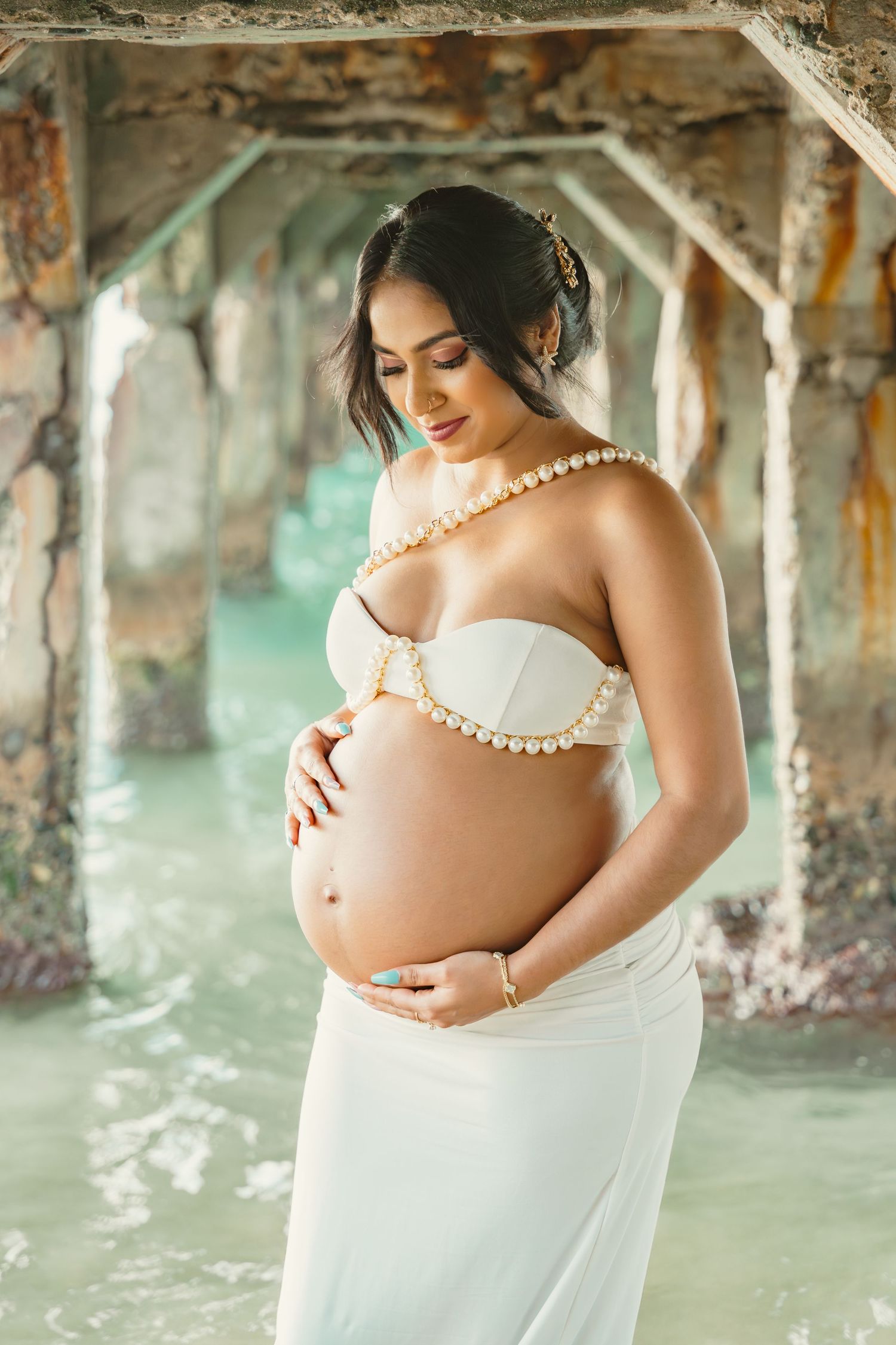 Pregnant woman in white pearl bikini top and skirt poses gracefully under pier by ocean.