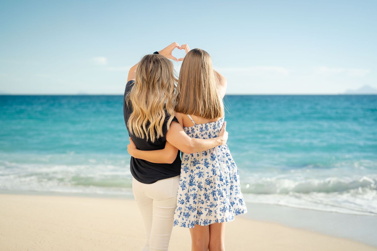 Two women making a heart shape with their hands while standing together on a sandy beach facing the ocean.