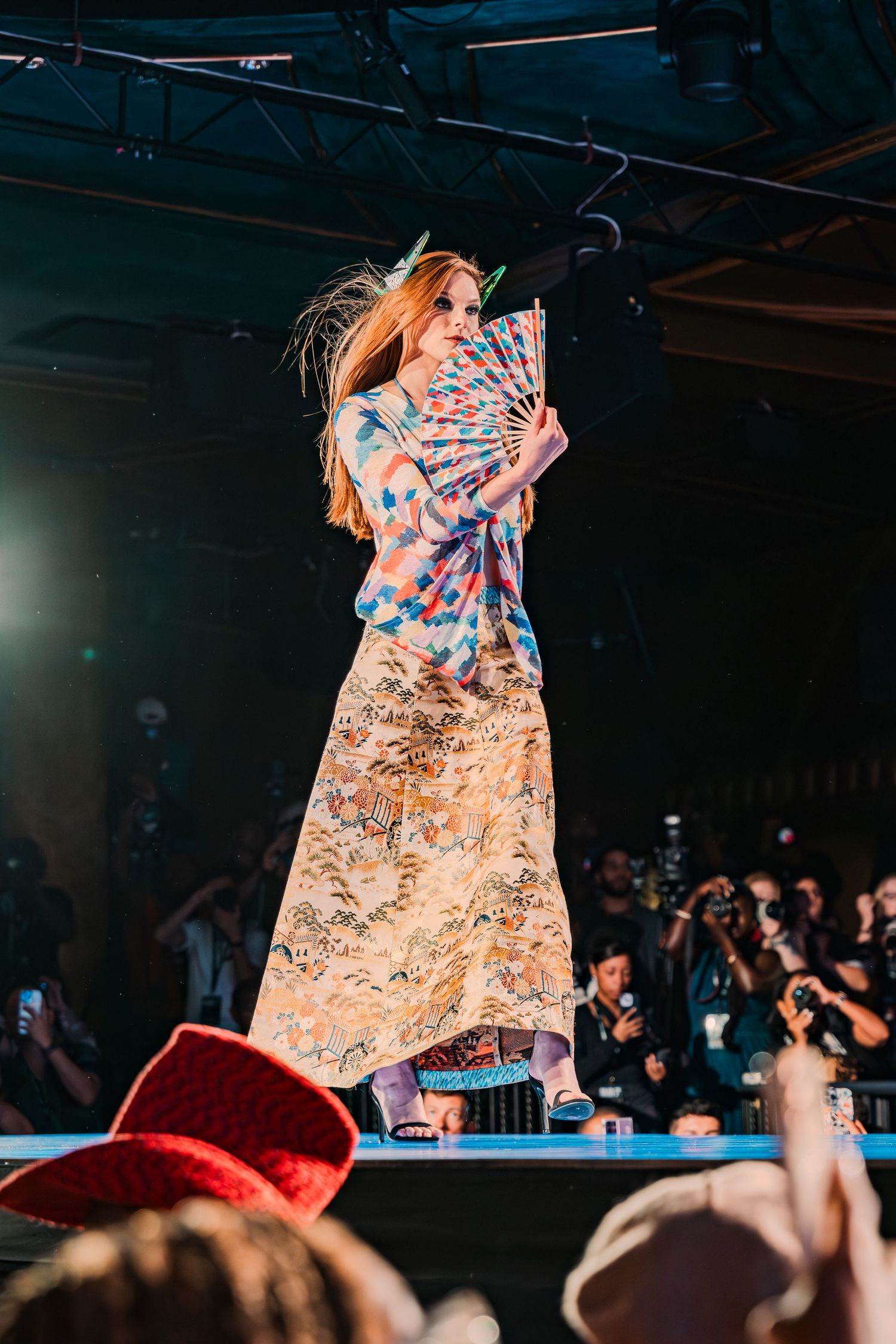 Model walks runway in floral kimono-style gown holding decorative fan at fashion show.